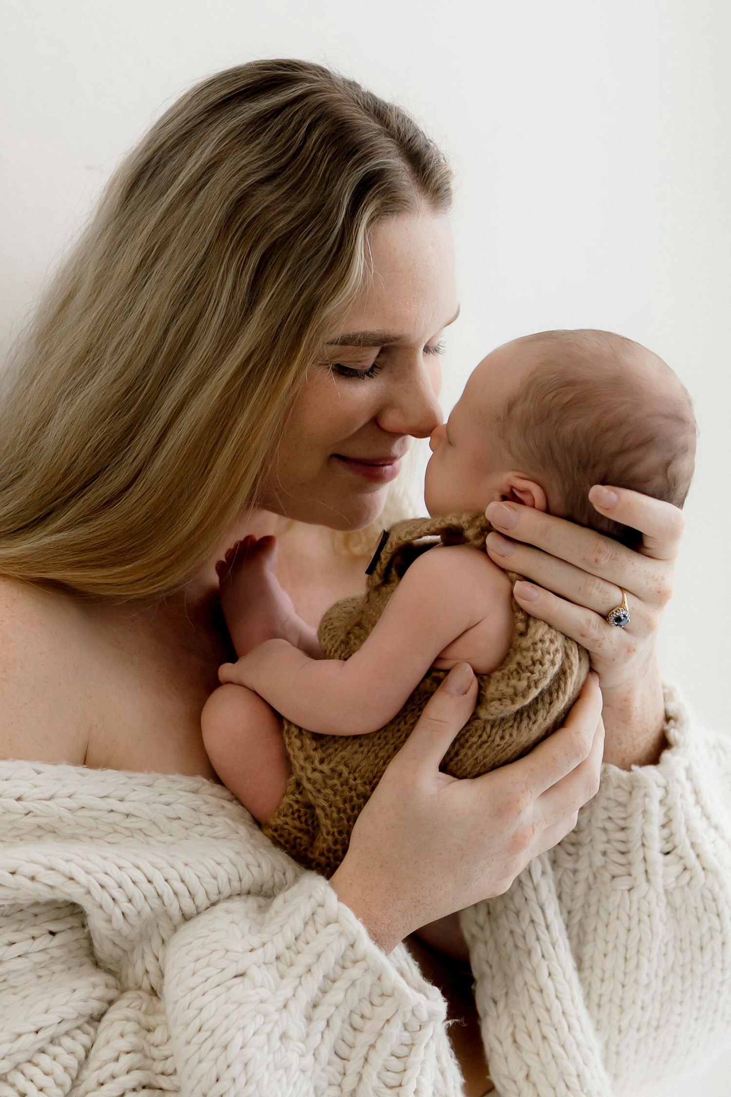 A mother tenderly holds her newborn baby wrapped in a brown knit blanket, sharing a gentle nose kiss.