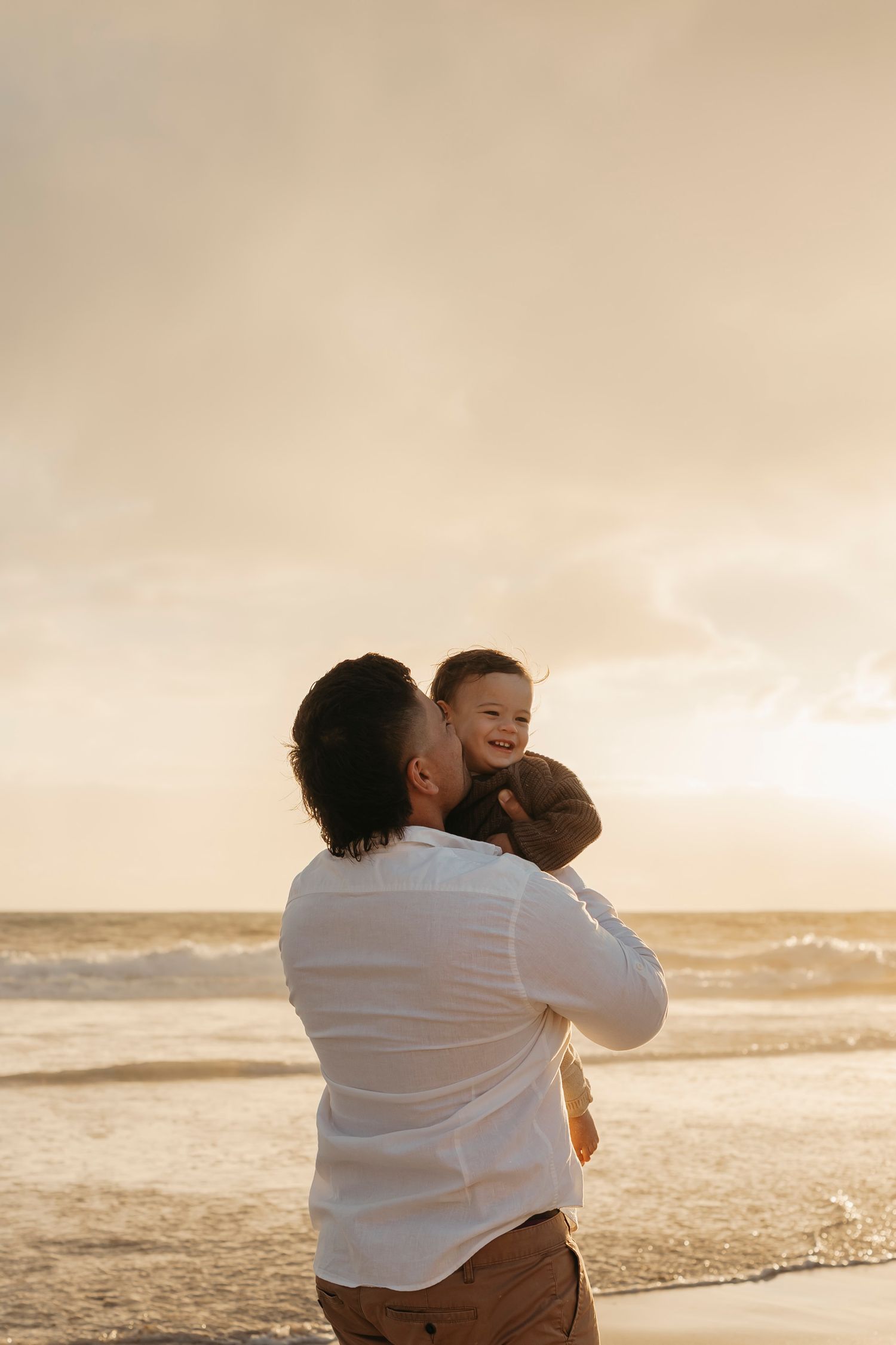 A parent and child share a tender moment at sunset on a beach with waves rolling in the background.