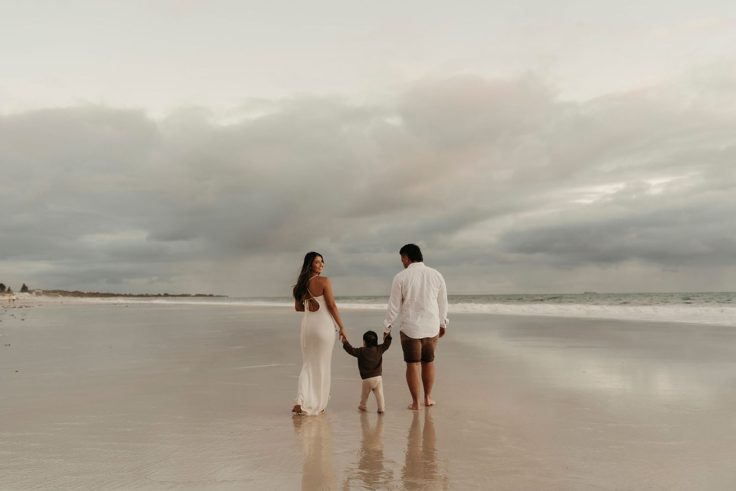 Small family walking hand in hand on an empty beach under cloudy skies.