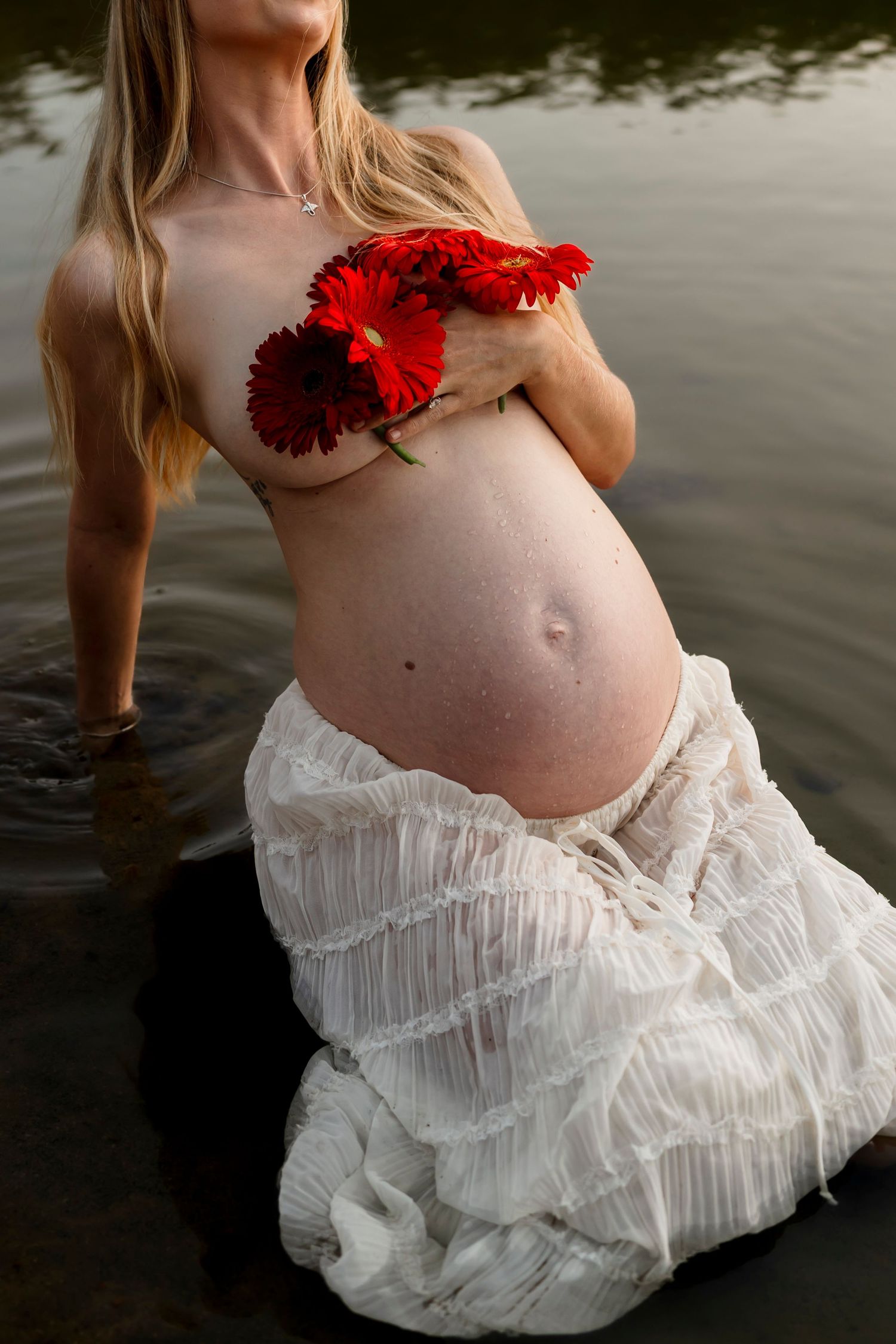 Artistic maternity photo showing a pregnant woman in a white dress standing in water holding red carnation flowers.