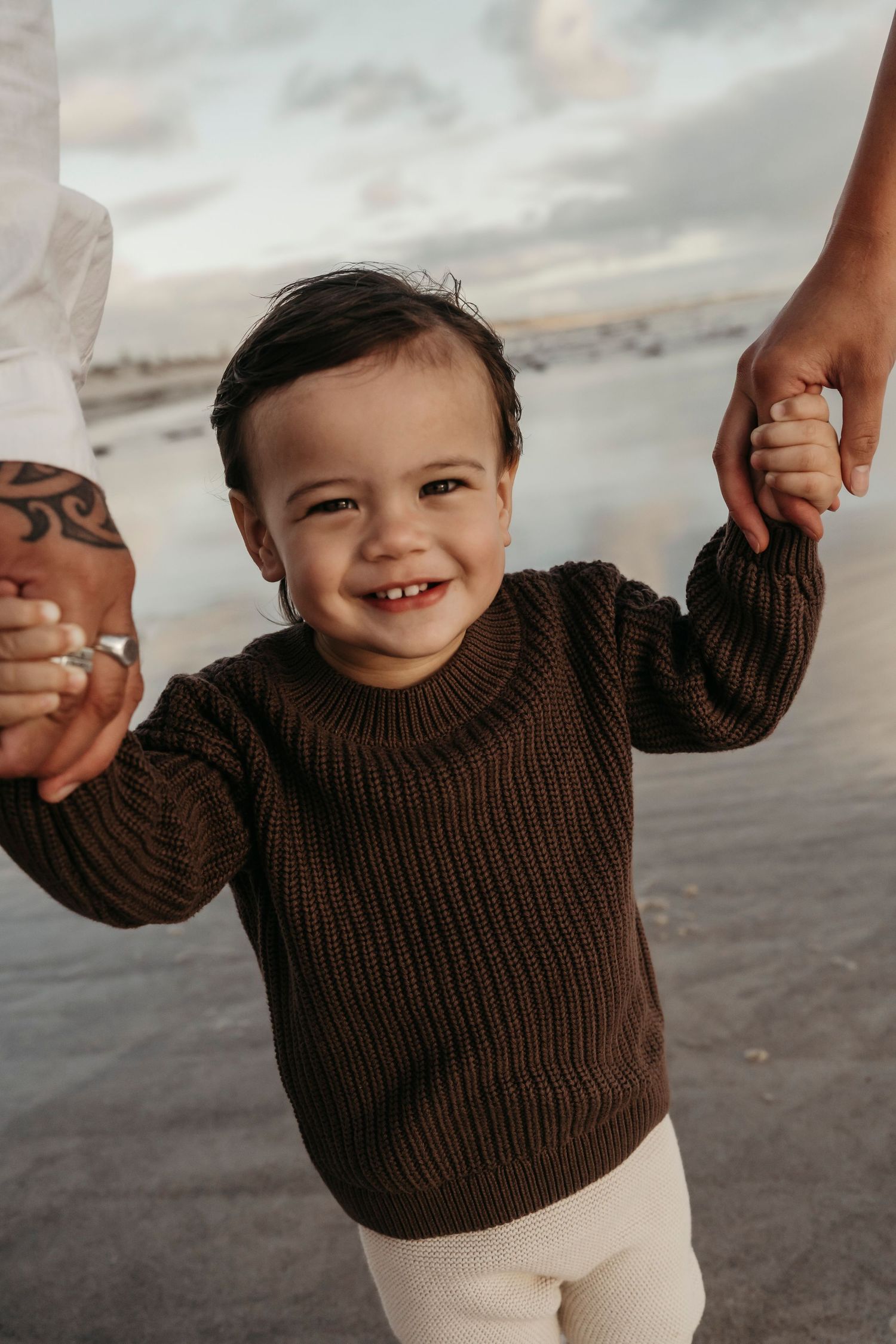 A small child in a brown sweater smiles while walking on the beach at sunset, holding hands with adults.