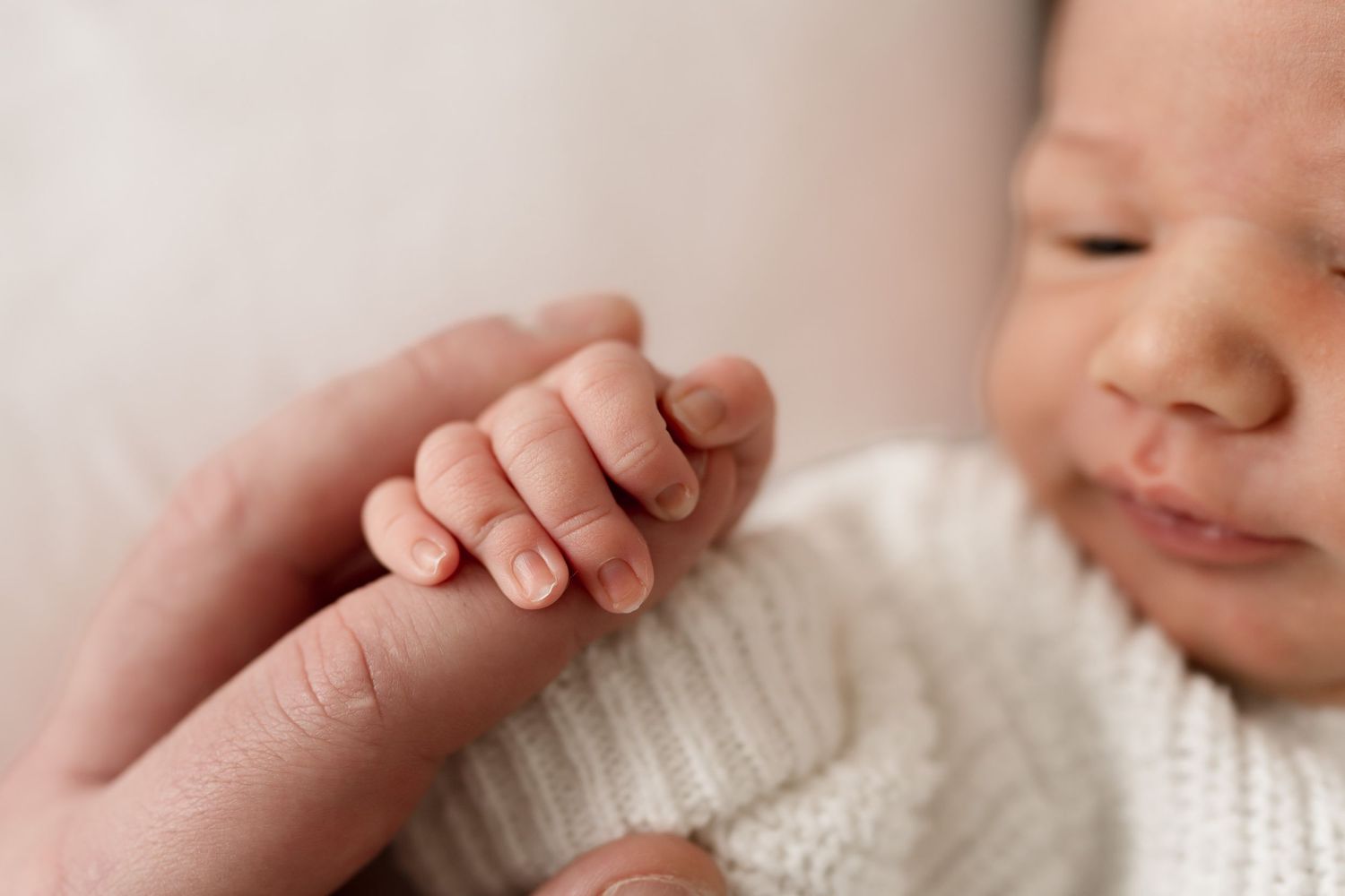 Tiny newborn hand grasping adult finger while wearing white knit sweater. Meg Rose Photos Perth WA.