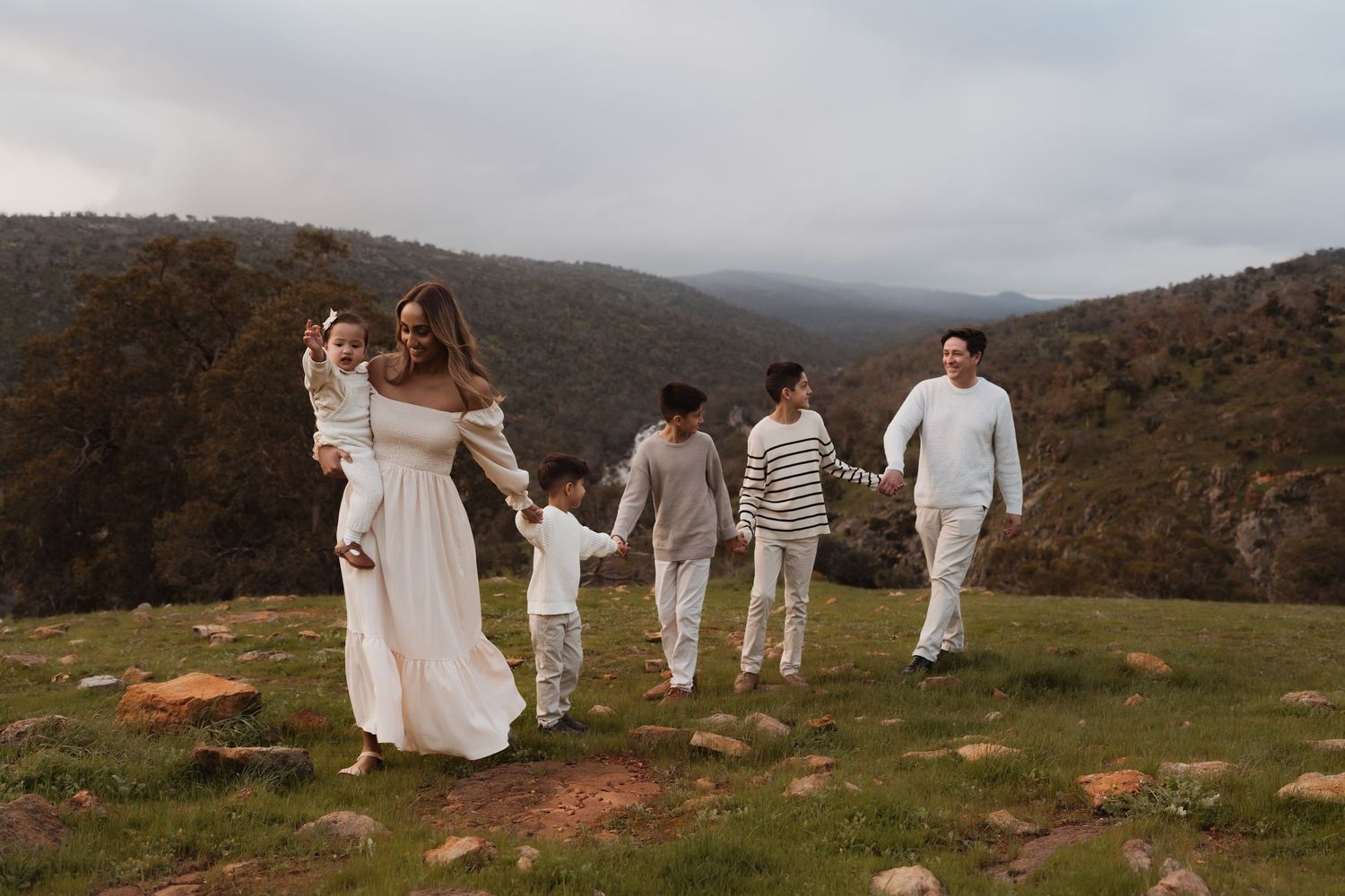A family wearing white outfits walks along a scenic mountain path under cloudy skies at dusk.