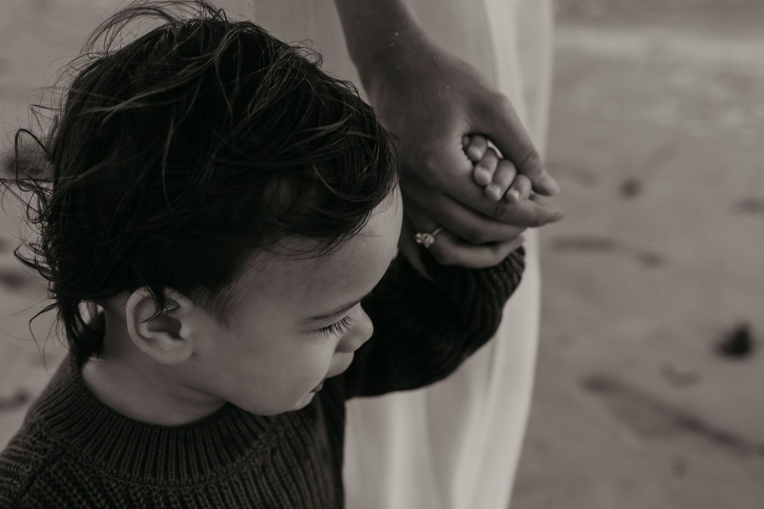 A black and white portrait of a small child in a dark sweater holding onto a parent's clothing.