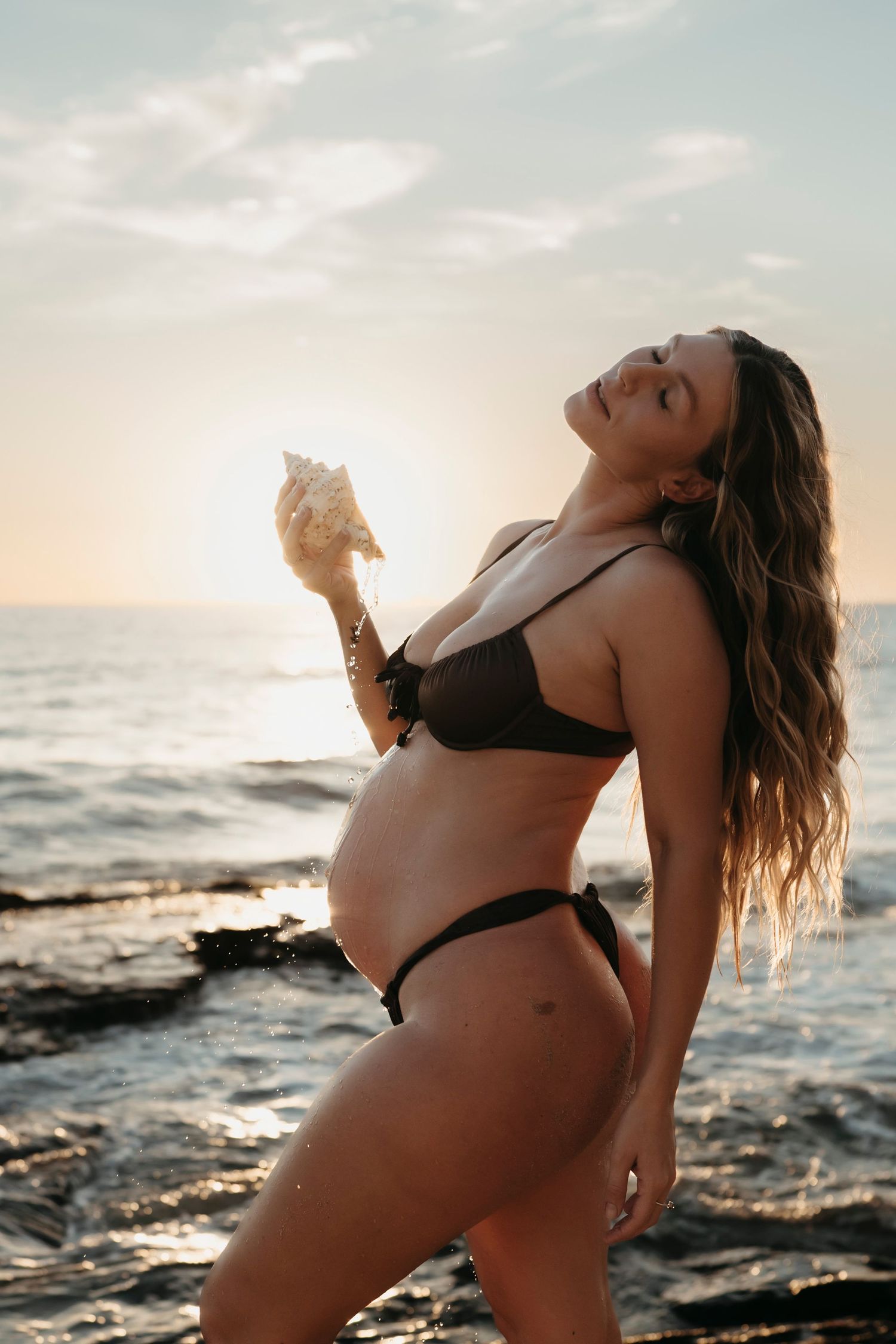 Woman in black bikini poses dramatically against ocean waves and sunset backdrop creating artistic silhouette.