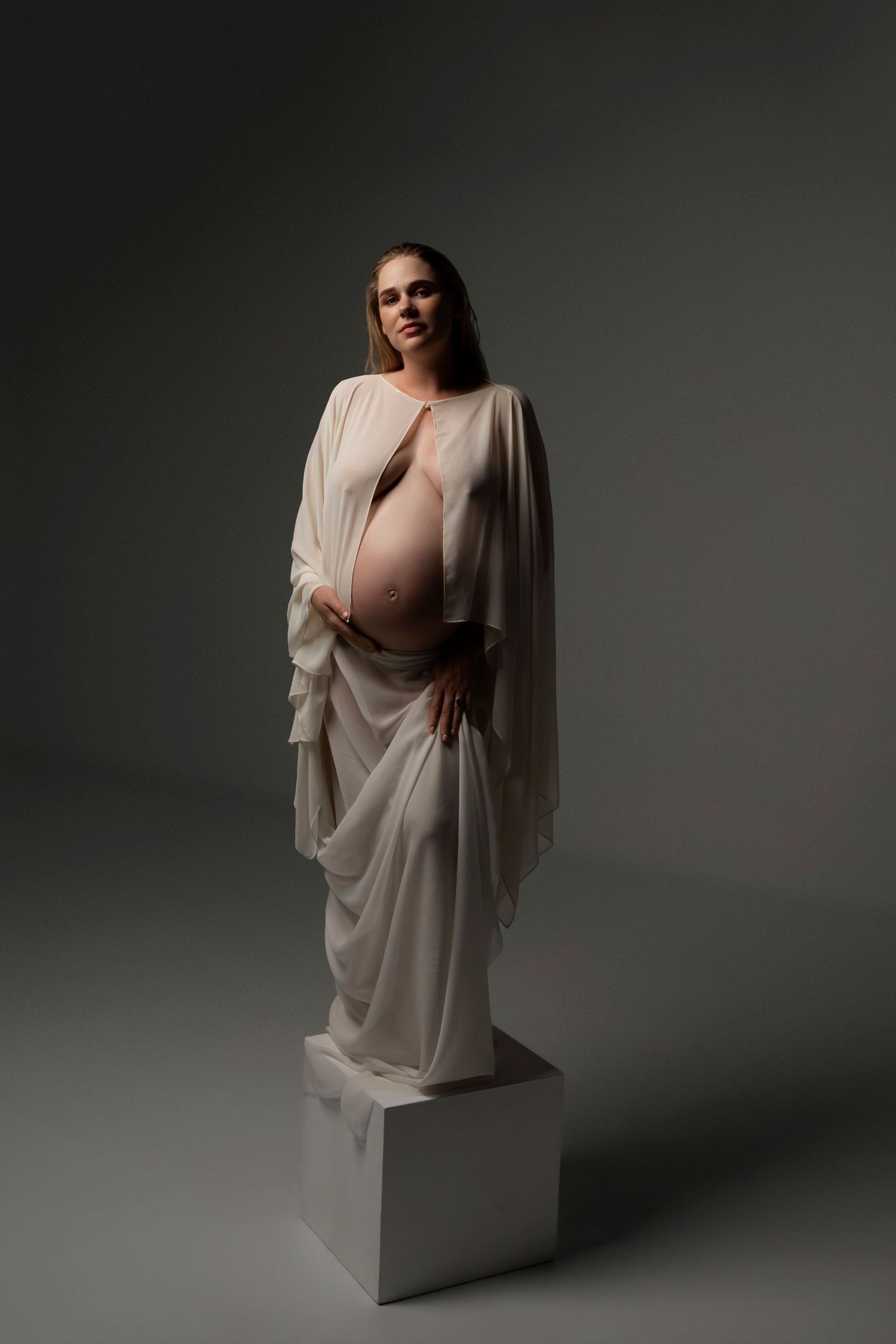 Pregnant woman in flowing white sheer drape gown standing on white cube pedestal against gray background.
