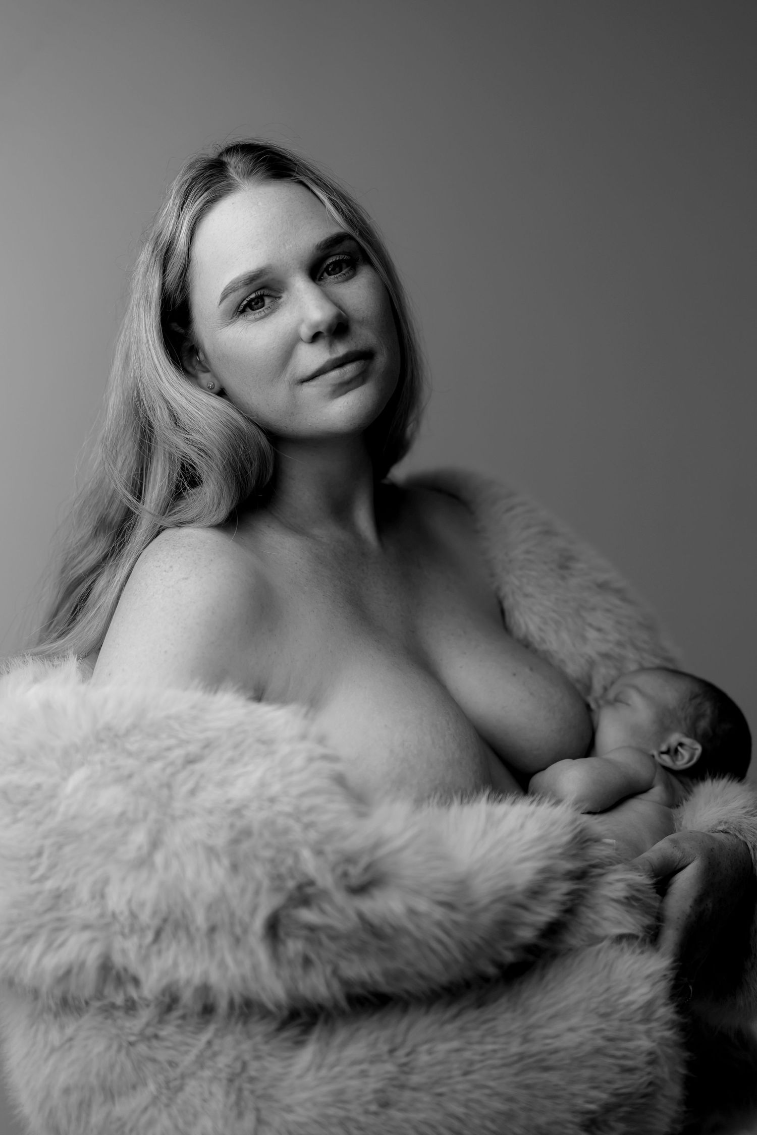 Black and white portrait of a woman breastfeeding her newborn baby, draped in a soft fur wrap.