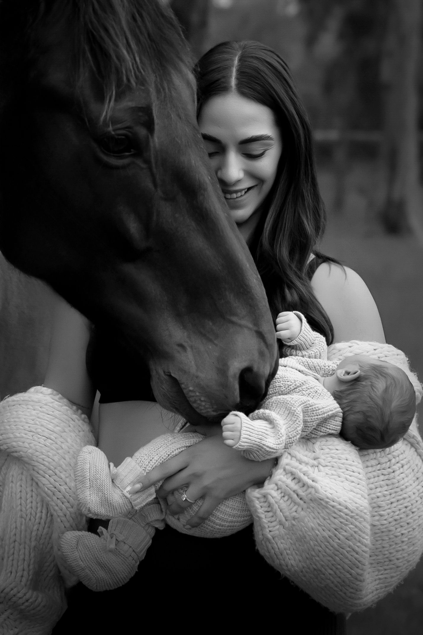 Black and white photo of smiling woman holding newborn baby while horse gently nuzzles infant.