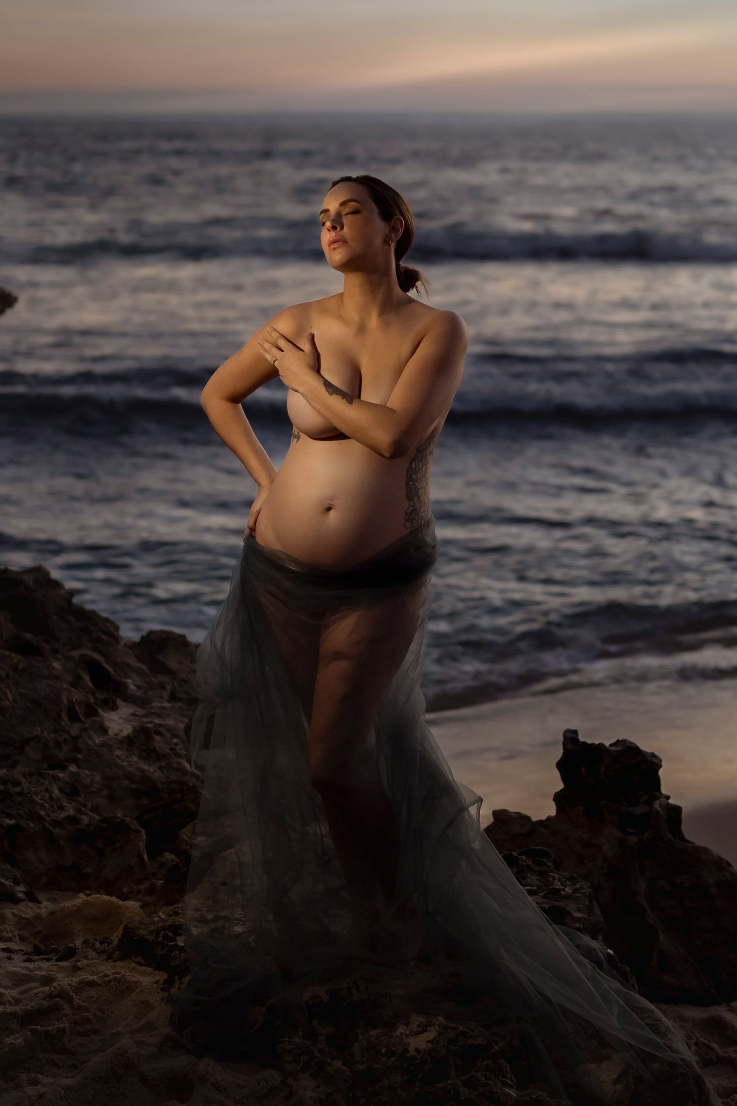 Pregnant woman in sheer teal skirt poses on rocky beach at sunset, cradling her baby bump near ocean waves.