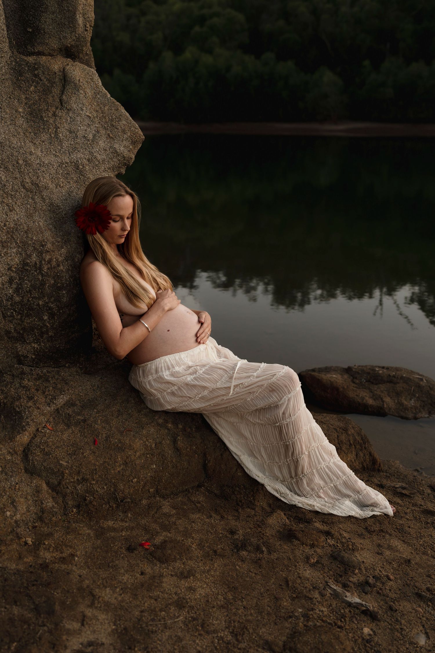 A tranquil maternity photo by a lake features an expectant mother in a white lace dress resting against a large rock.