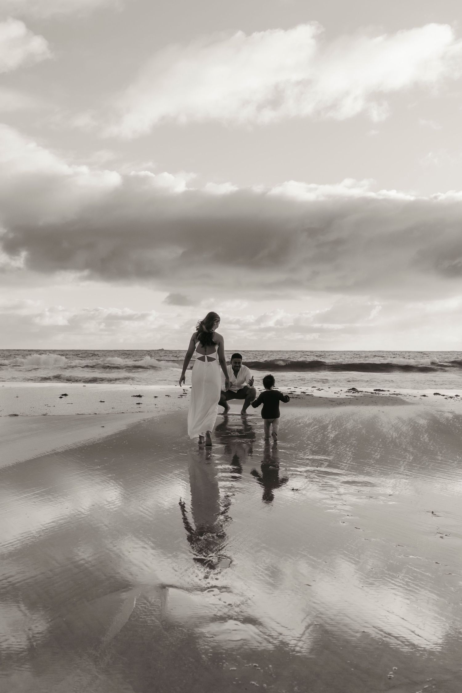Black and white image of three silhouettes walking along a reflective beach shoreline at dusk.