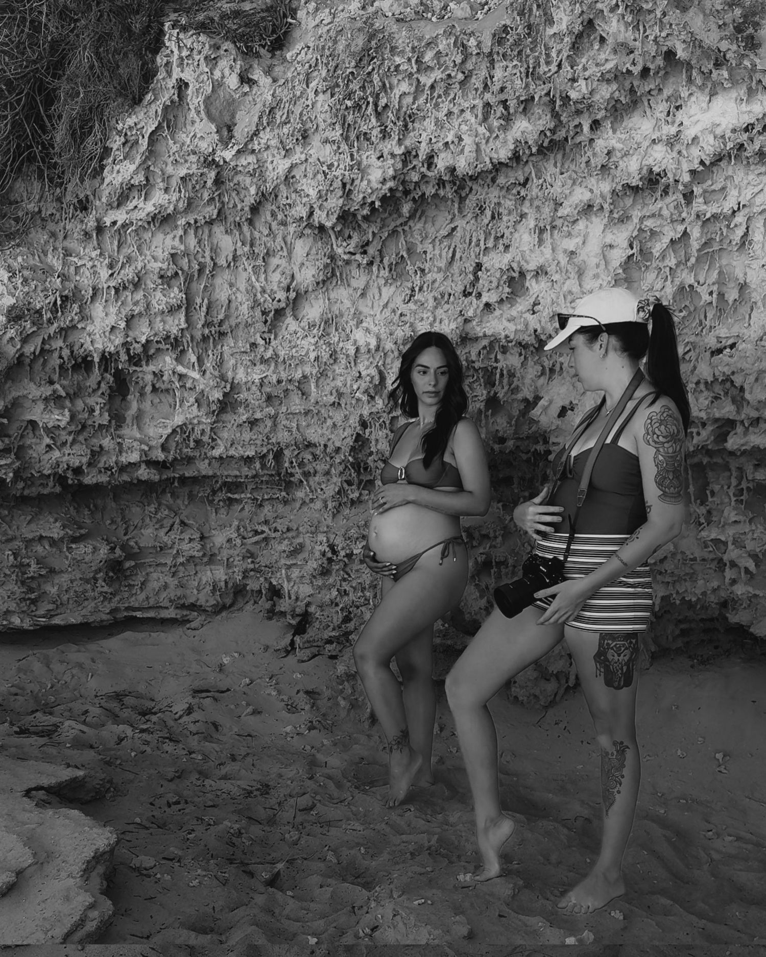 Two pregnant women in swimwear pose together on a sandy beach beside a rocky cliff wall in black and white.