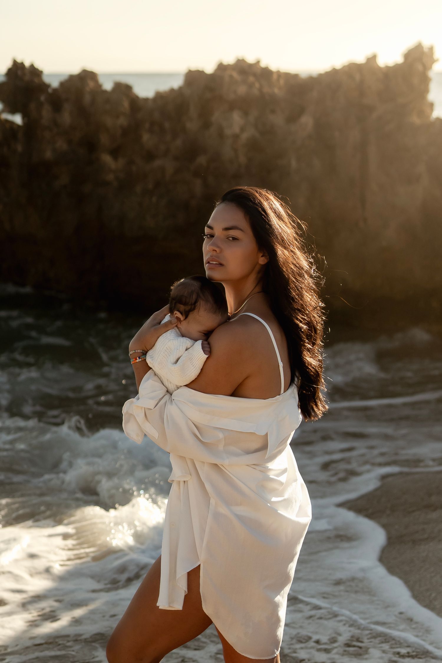 A woman in a white dress holds a newborn baby on a rocky beach at golden hour sunset.