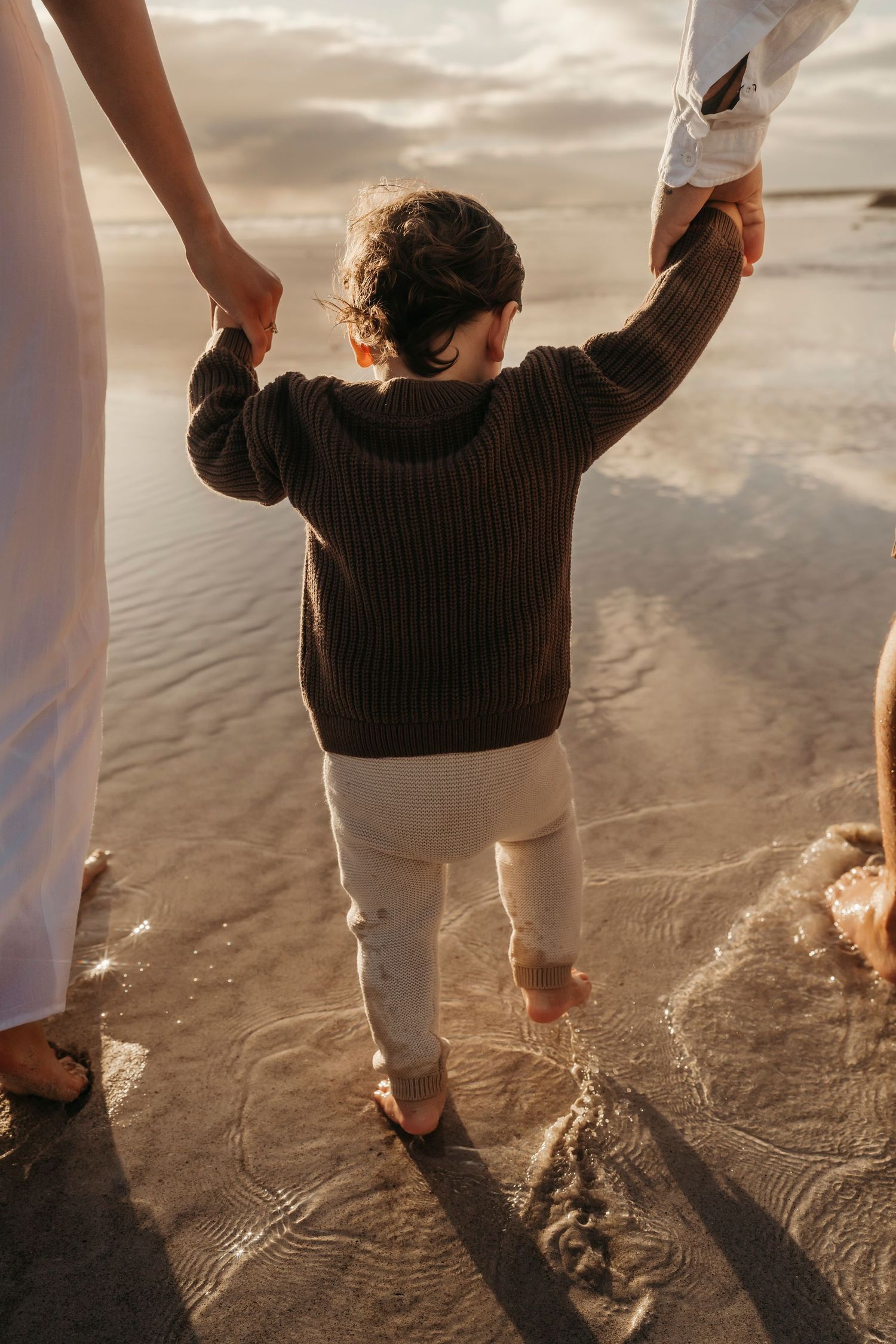 A small child in a brown sweater holds hands with adults while walking barefoot on a sandy beach at sunset.