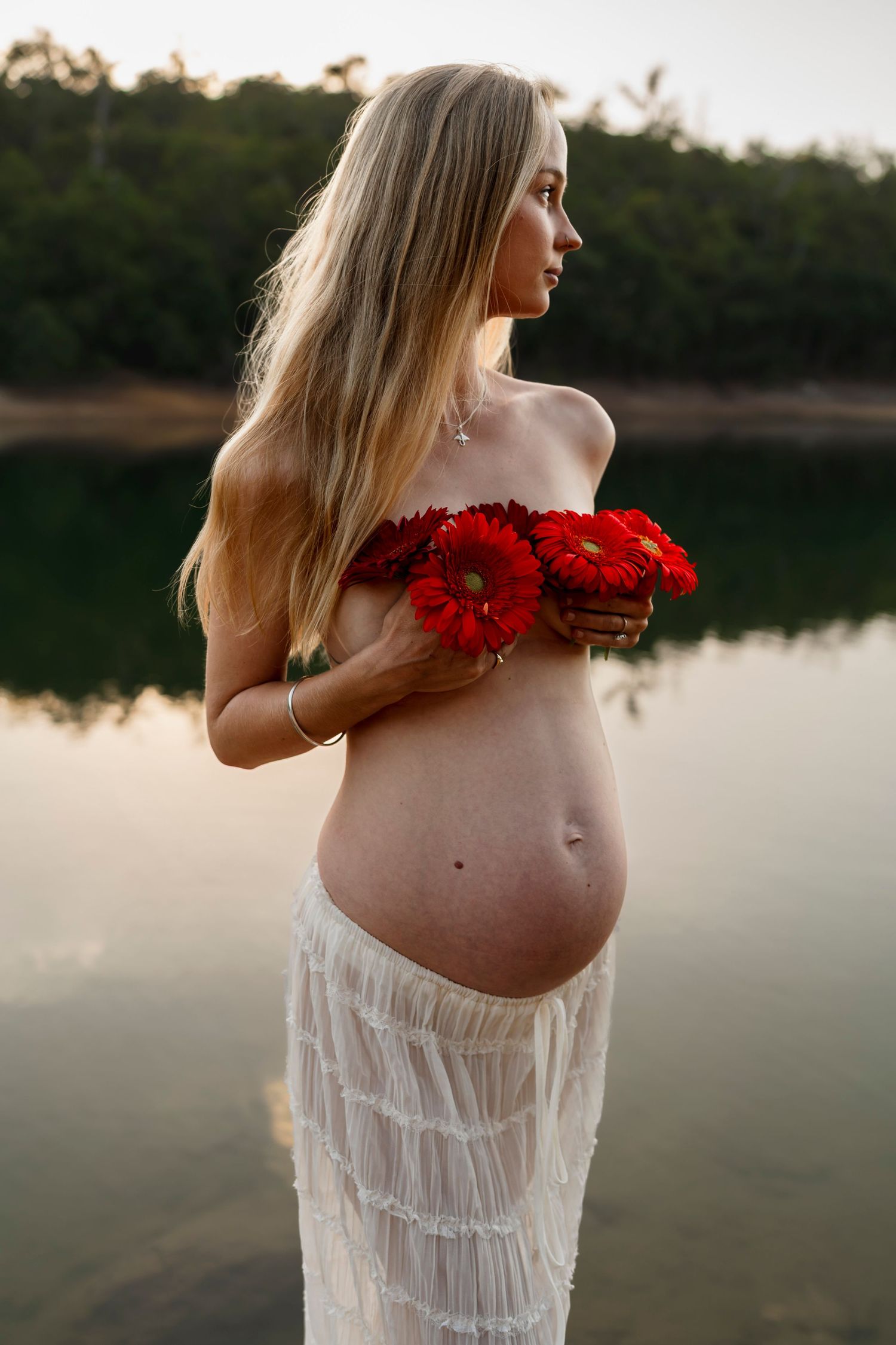 Expectant mother in white dress holding red flowers stands by serene lake at sunset.