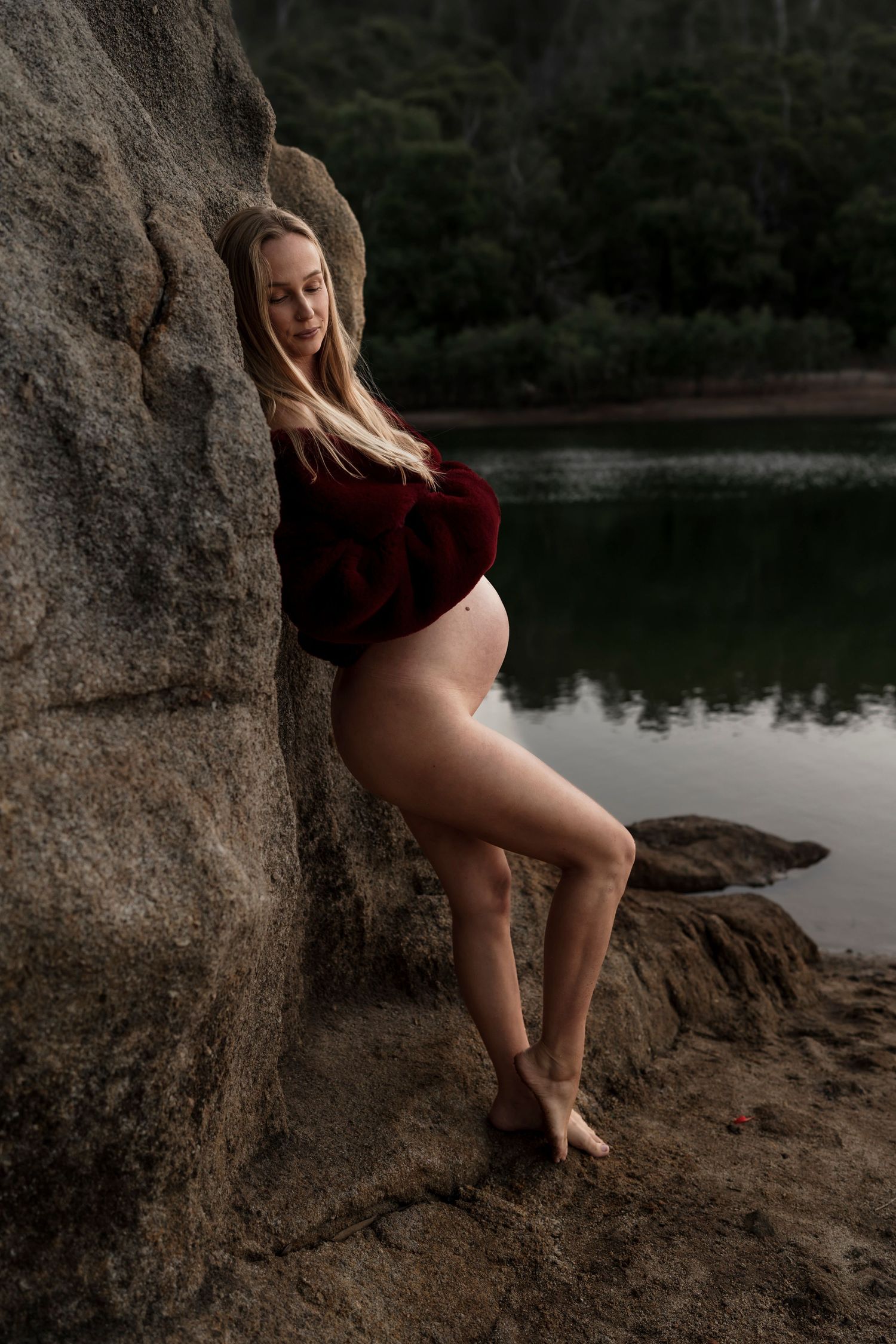 Maternity portrait in a red velvet bodysuit against rugged rocks by a serene lake.