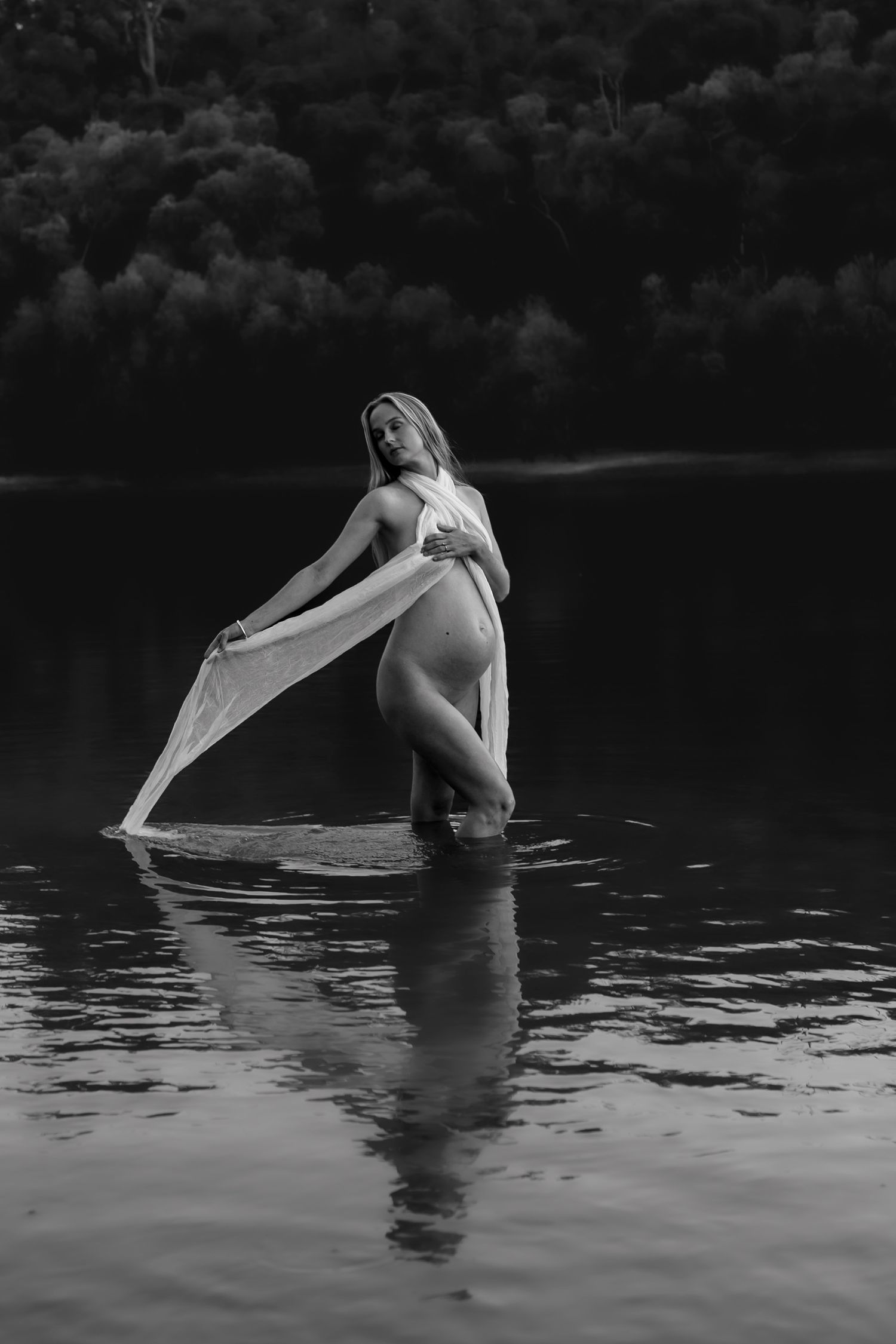 Artistic black and white portrait of an expecting mother standing in tranquil water while sunlight illuminates her flowing dress.