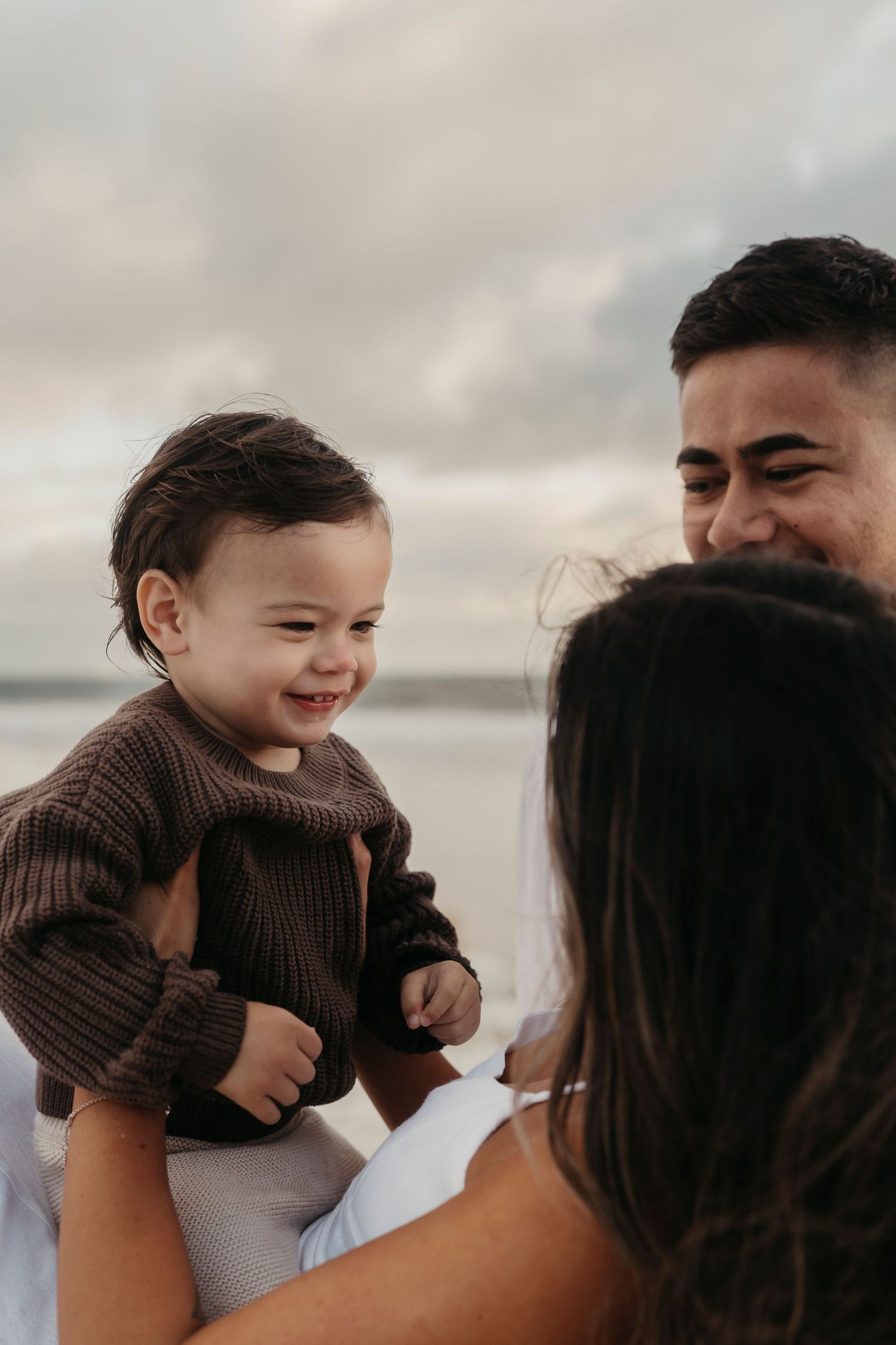 A joyful candid family moment captured outdoors with a small toddler in a brown sweater against a cloudy sky.
