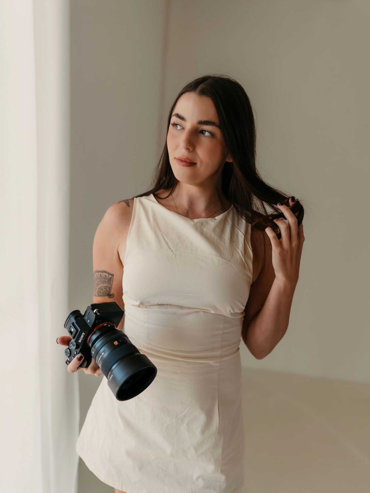 A photographer wearing a white tank top holds a camera while standing against a neutral wall in a series of poses.