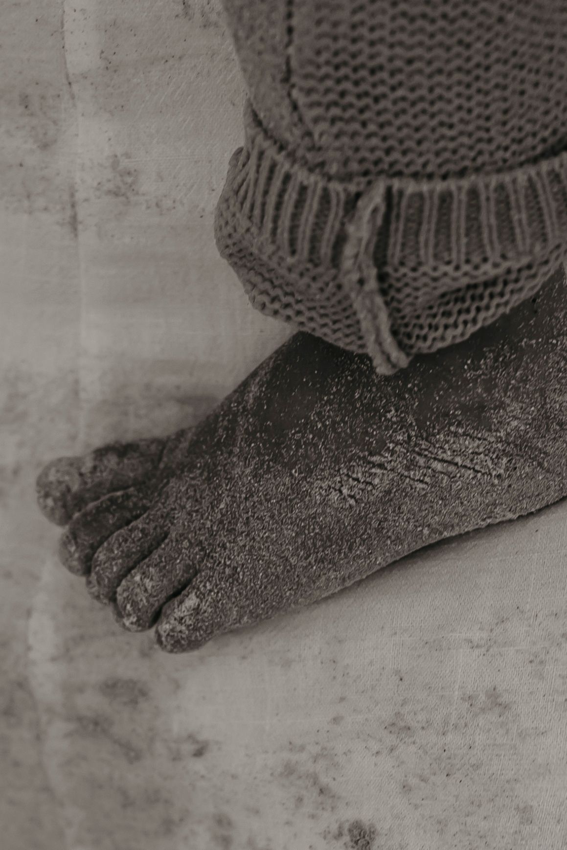 Black and white close-up of dirty feet wearing a knitted sock, showing gritty texture against a concrete floor.