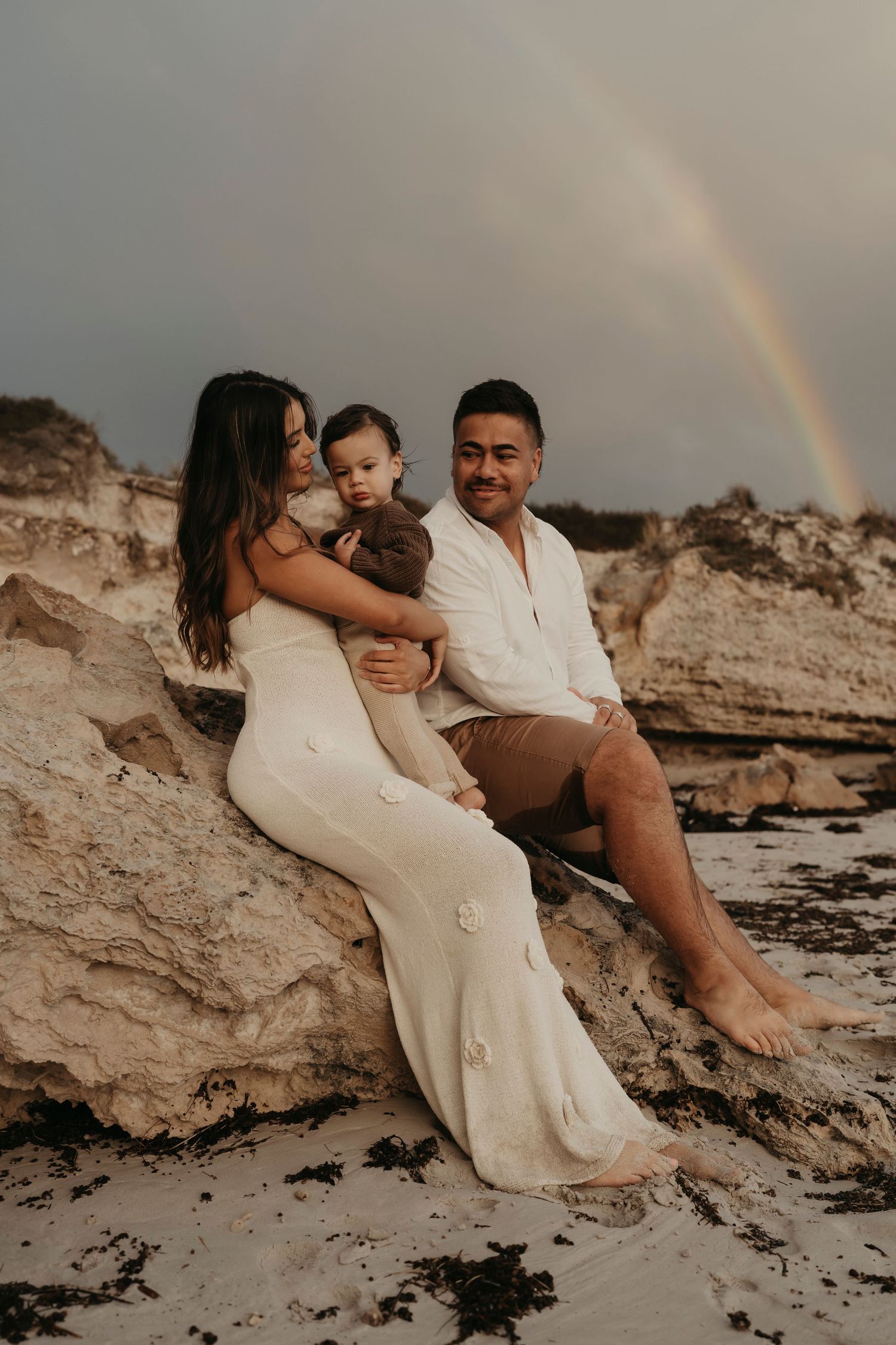 A family sits together on beach rocks at sunset with a rainbow visible in the moody sky.