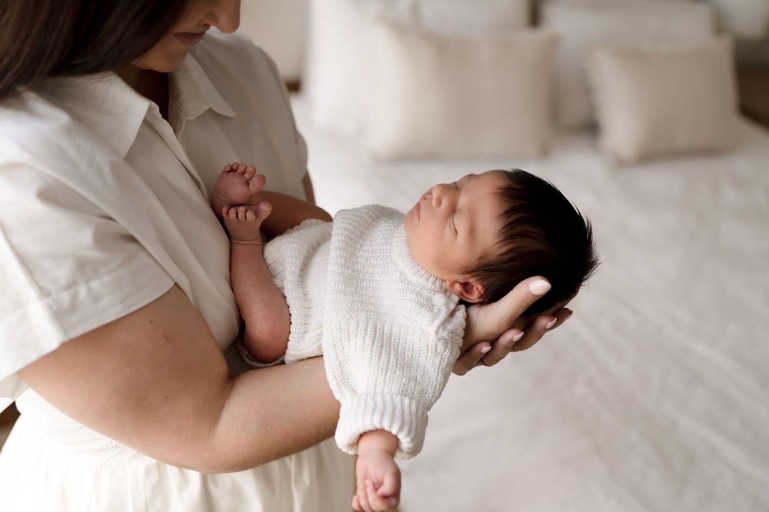 Parent cradling peaceful sleeping newborn baby dressed in white knit outfit on white bedding.