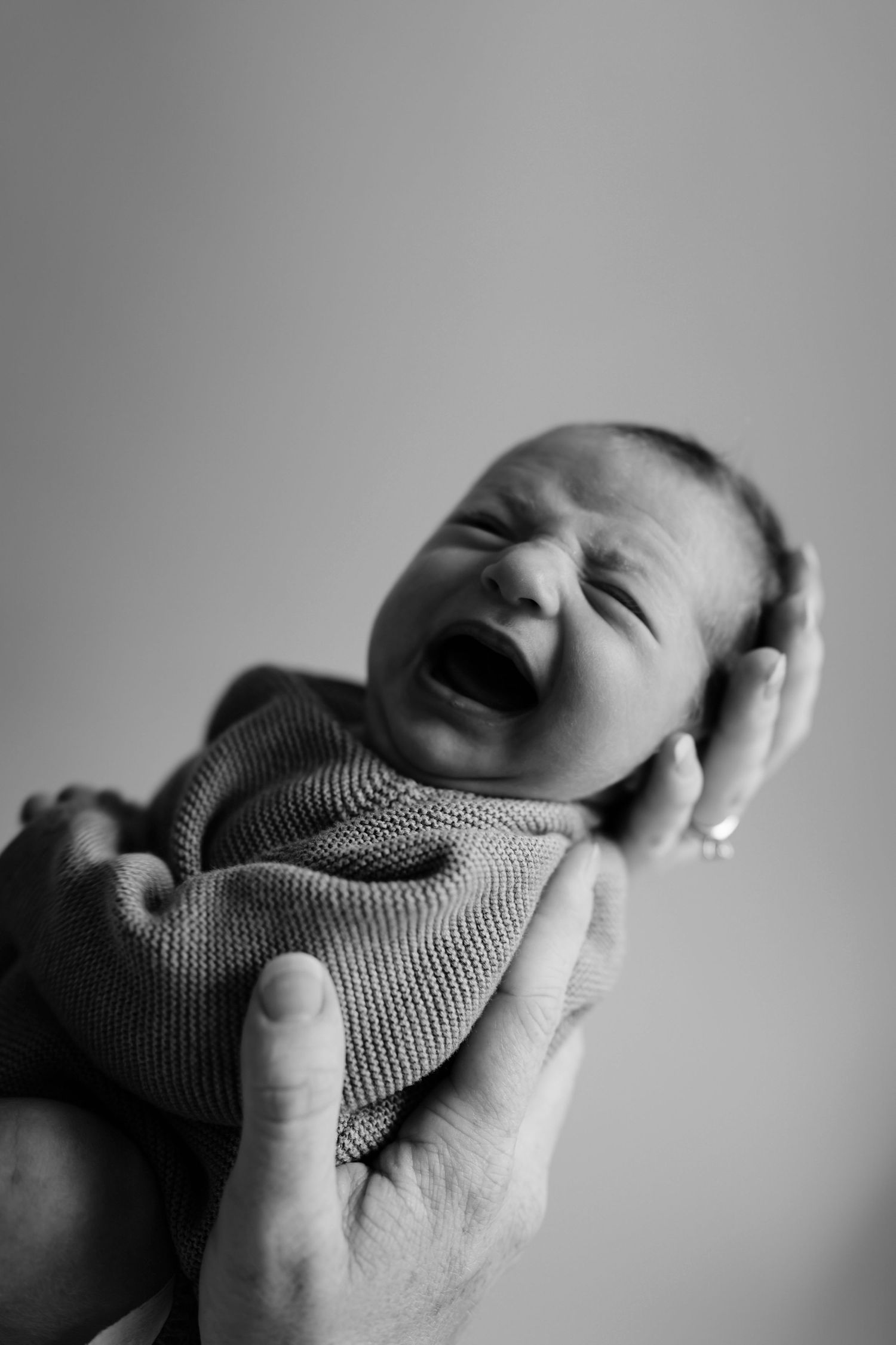 Black and white photo of a crying newborn baby being cradled in adult hands wearing a knit outfit.