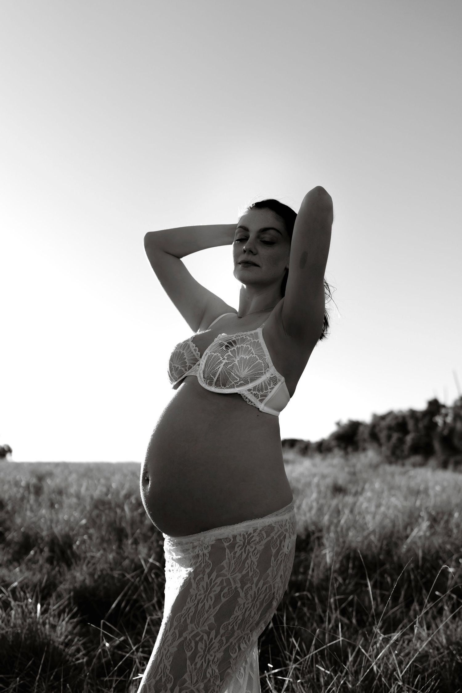 Black and white photo of a pregnant woman in lace lingerie posing gracefully in a field.