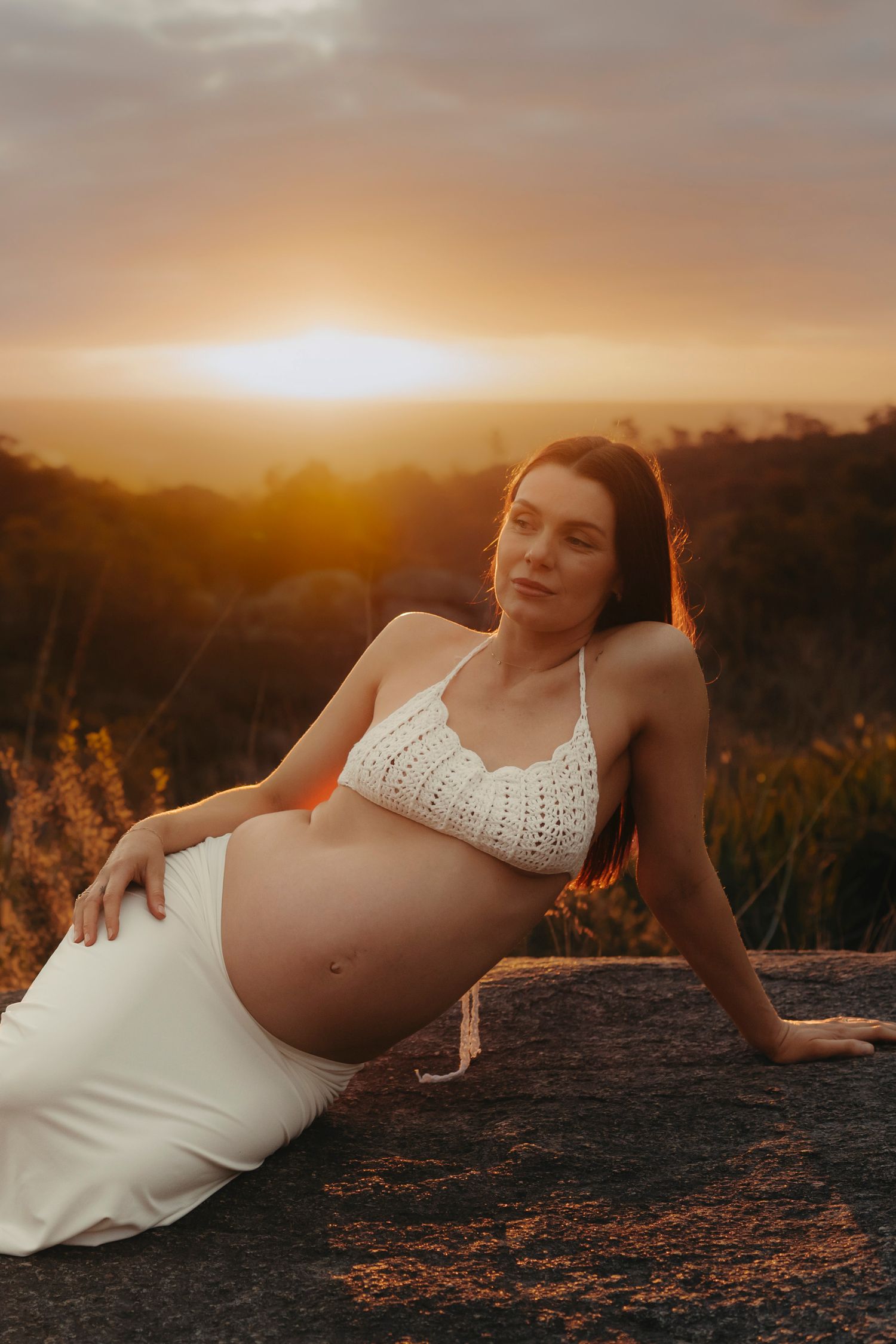 A pregnant woman in a white dress poses at sunset during a maternity photoshoot in a golden field.
