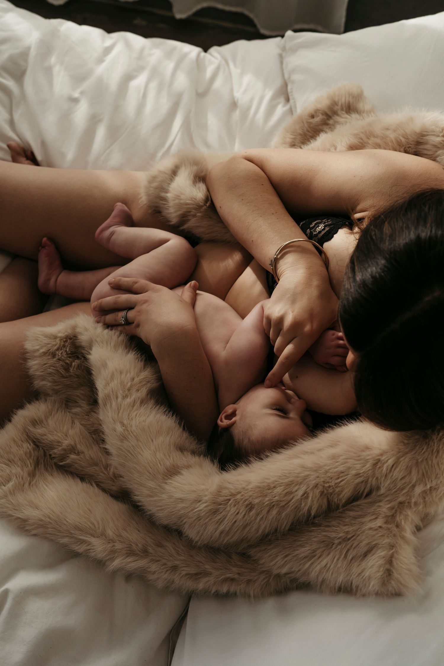 Mother breastfeeding newborn baby while lying on white bed wrapped in cozy faux fur blanket.