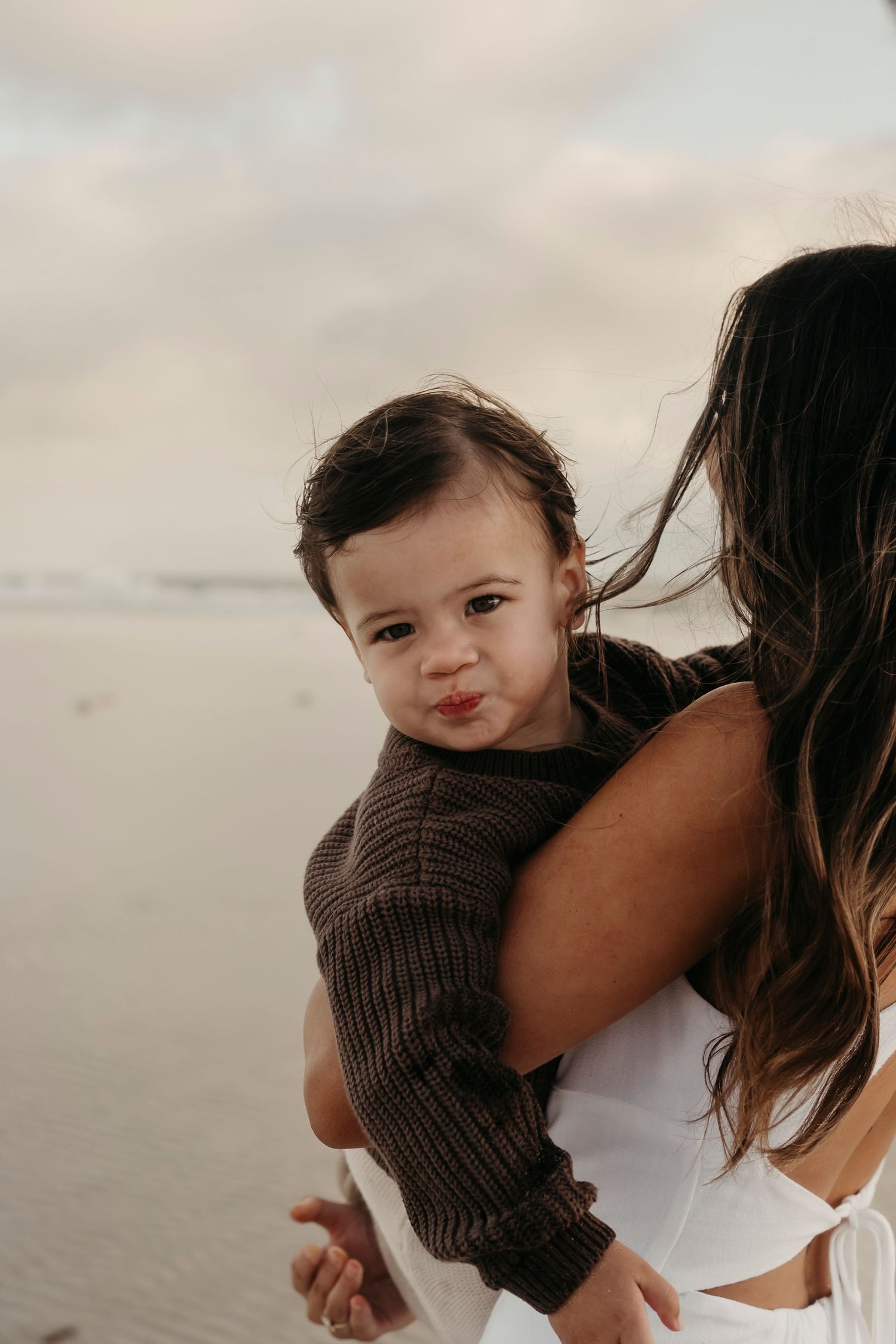A small child wearing a brown sweater looks over their caregiver's shoulder at the beach during sunset.