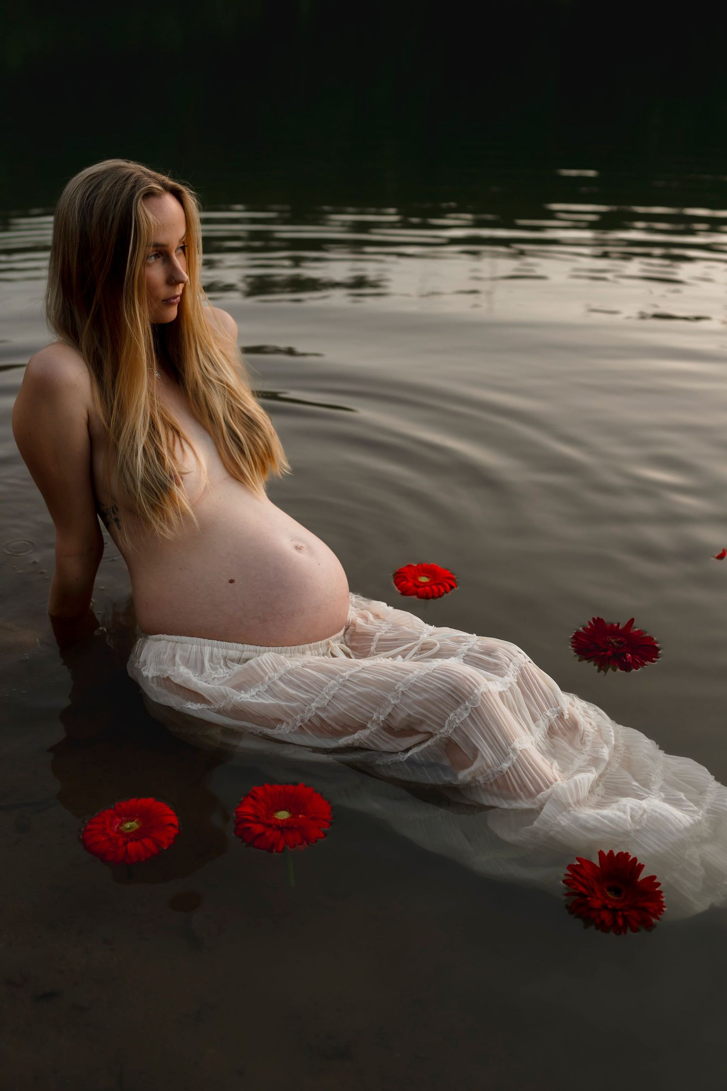 Artistic maternity portrait in water with flowing white fabric and floating red gerbera daisies creating ethereal atmosphere.