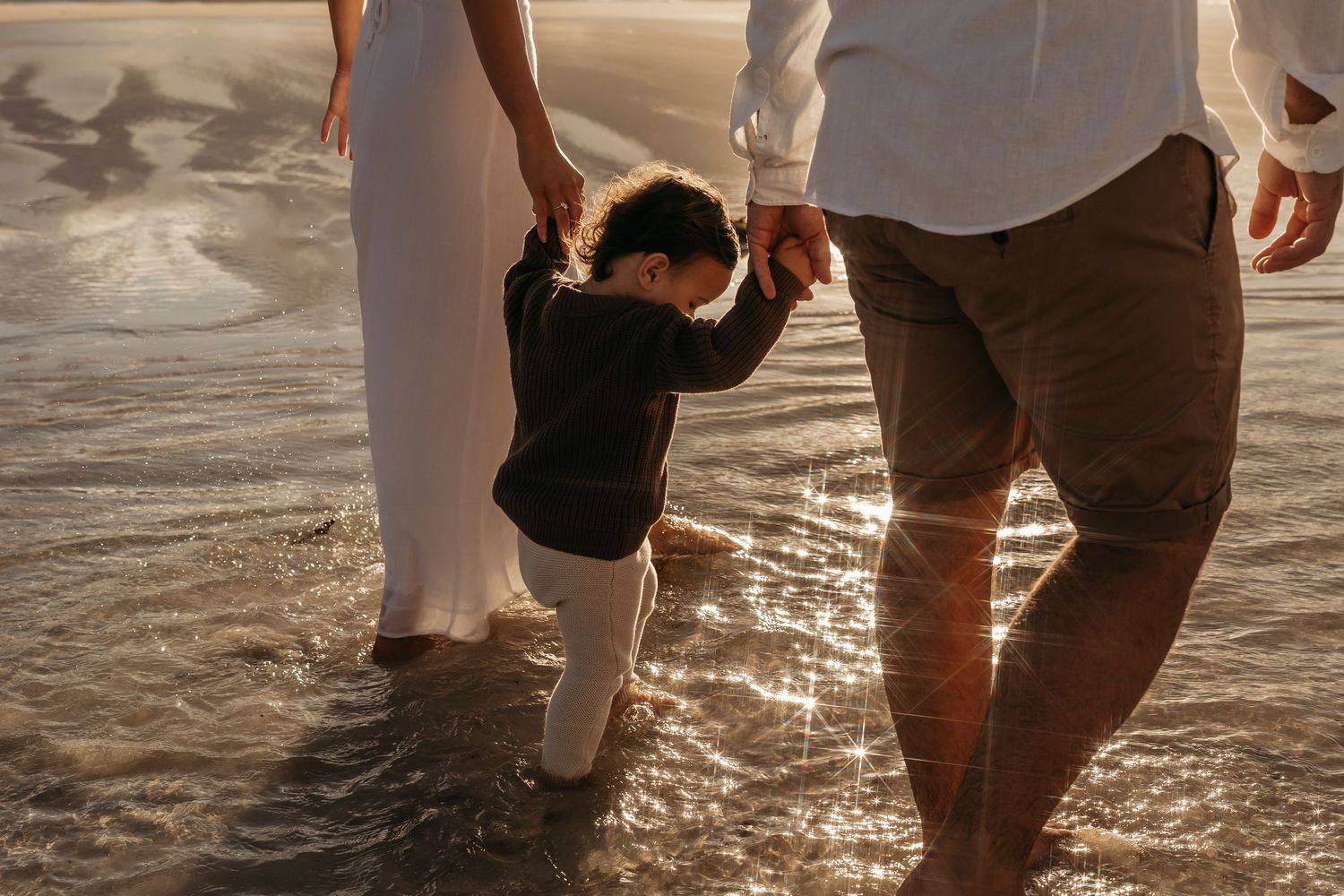 Small child holding hands with parents while walking on beach at sunset with golden light reflecting off wet sand.