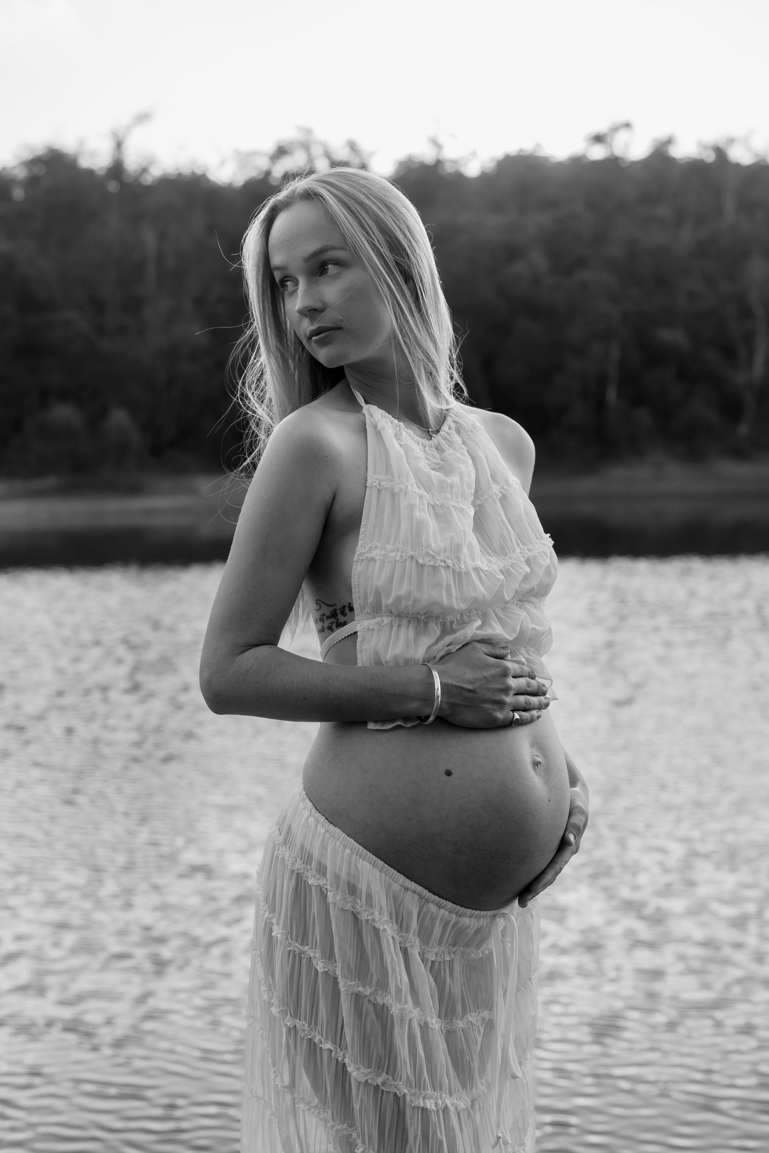 Black and white maternity portrait by a lake showing a pregnant woman in a flowing white tiered dress.