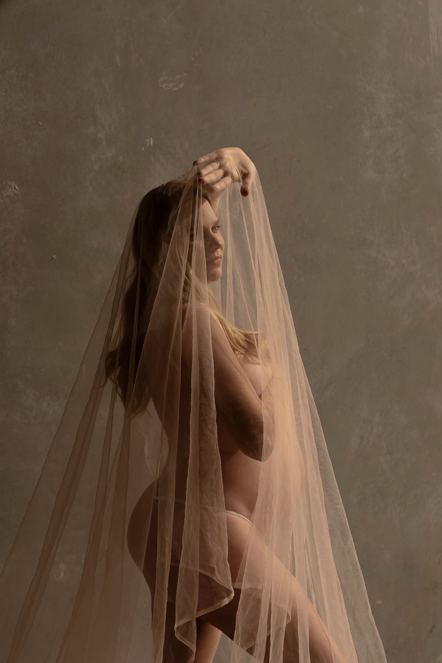 A woman draped in sheer tulle veil poses gracefully against a muted green backdrop in an artistic studio portrait.