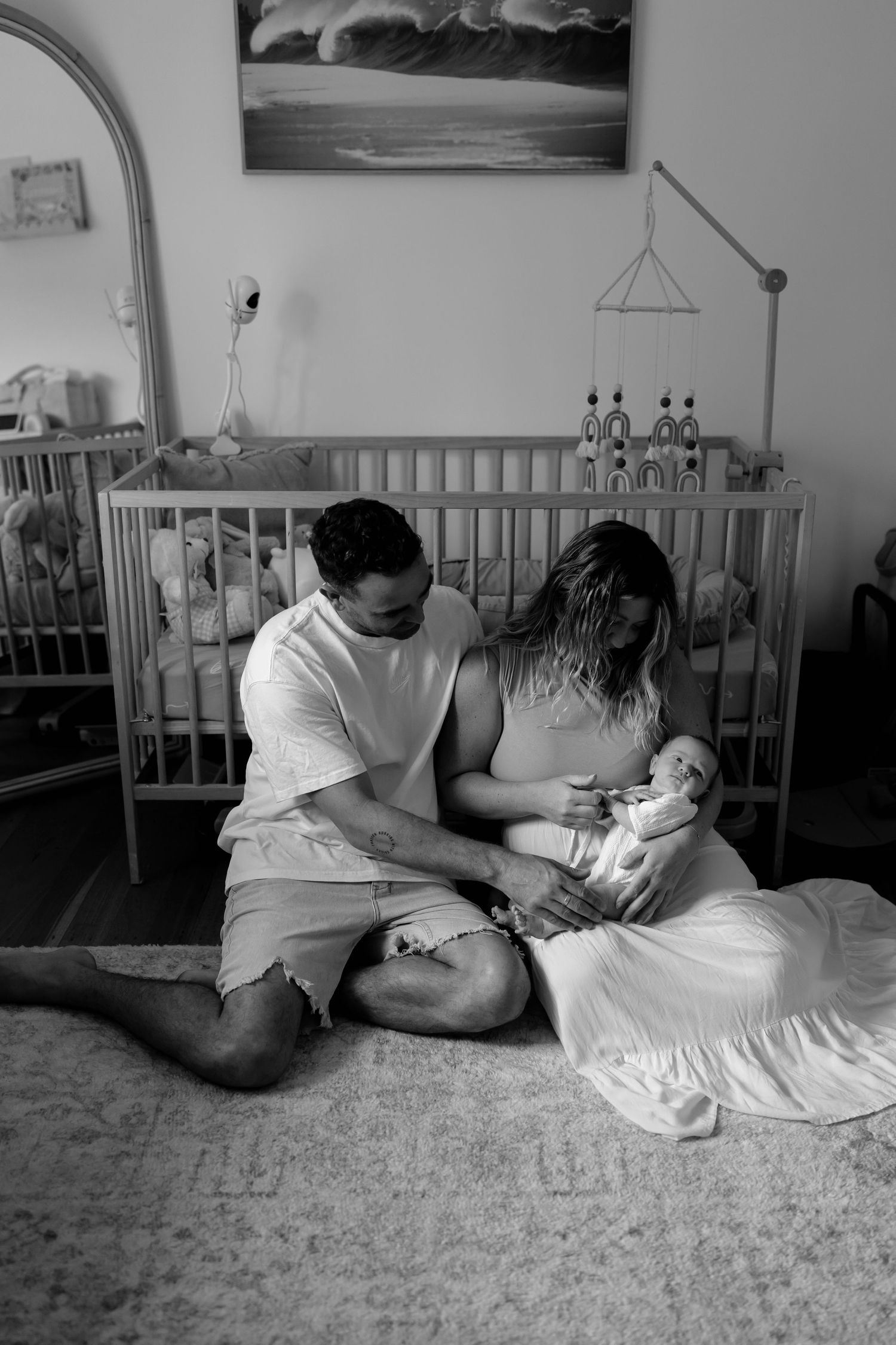 Black and white photo of parents sitting on nursery floor with newborn baby in front of crib.