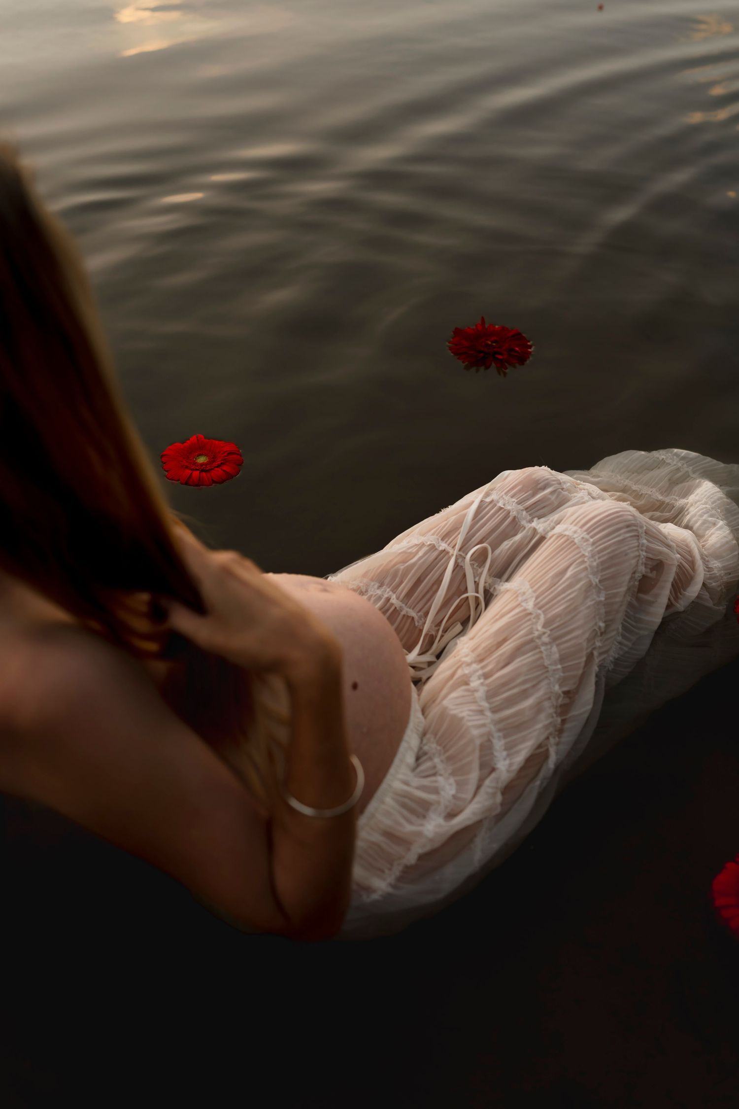 Artistic photo of a flowy white dress flowing in dark water with red flowers floating nearby.