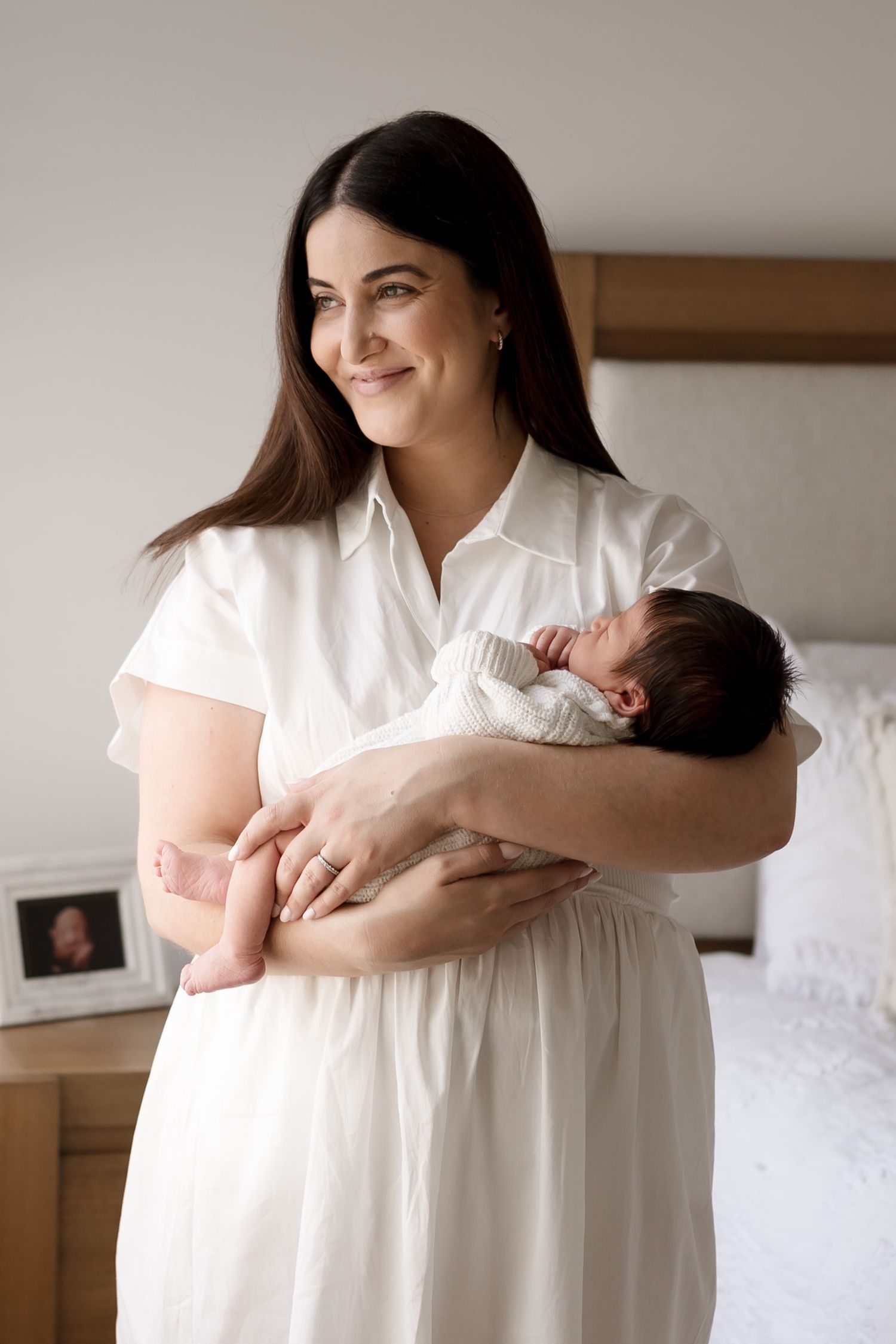 Woman in white dress holding sleeping newborn baby in minimalist bedroom setting. Meg Rose Photos Perth WA.
