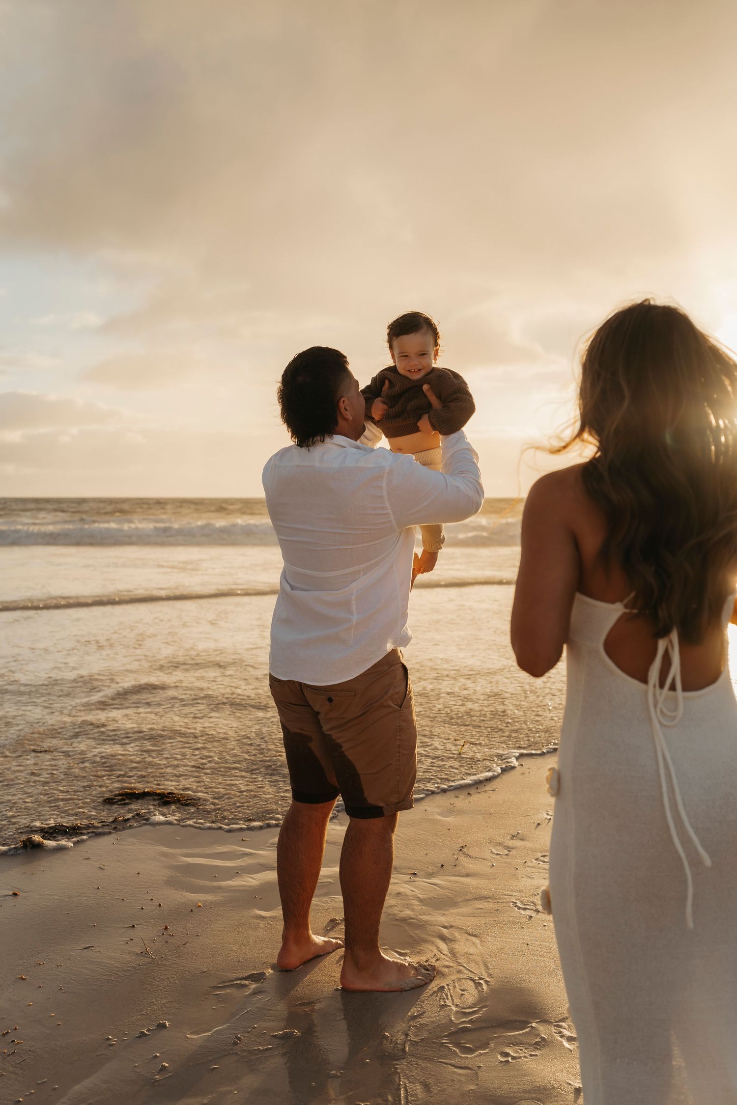 A couple enjoying sunset at the beach with a toddler against a golden sky and ocean backdrop.