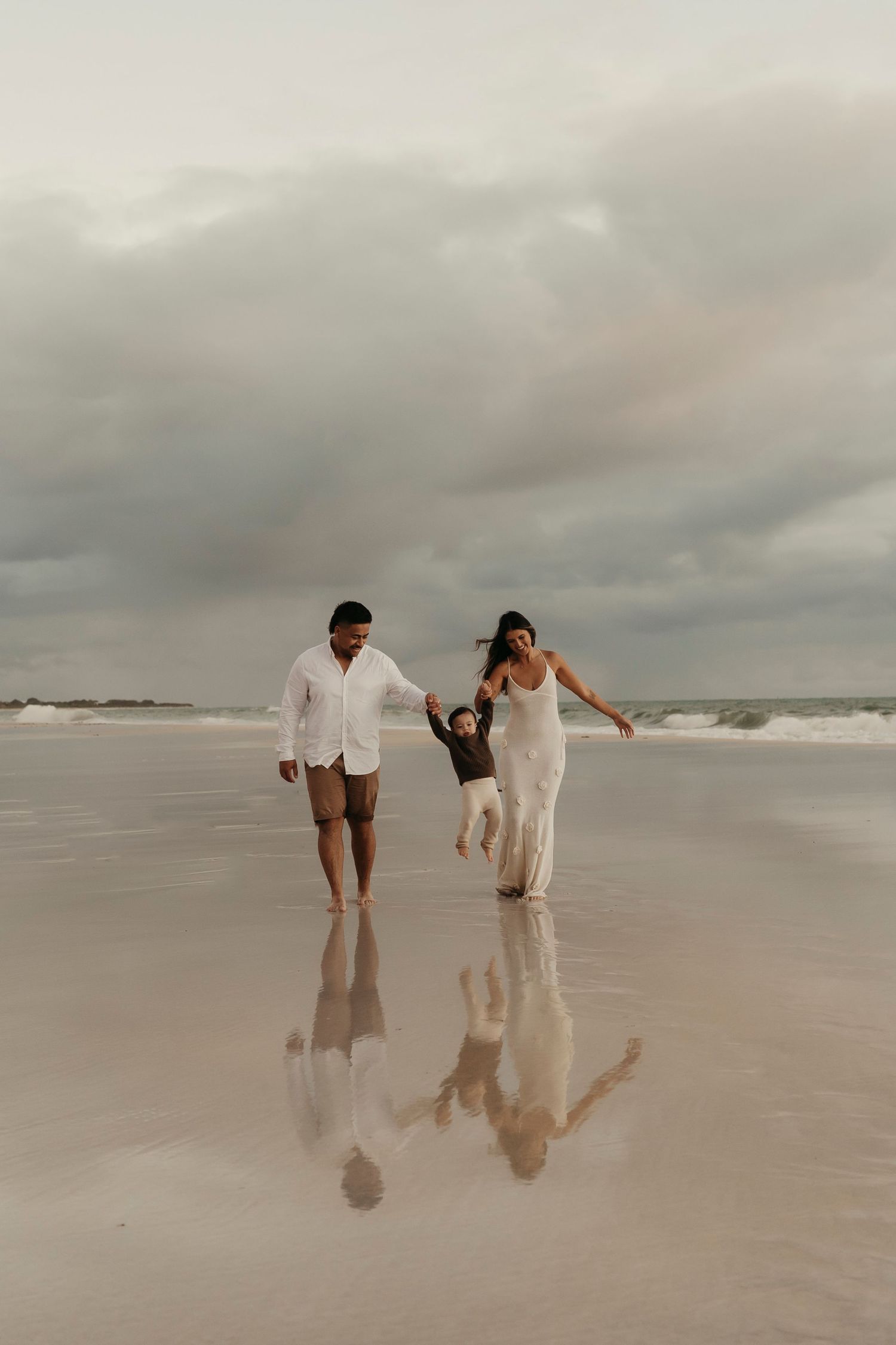 Family walks together holding hands on a misty beach with their reflections visible in the wet sand.