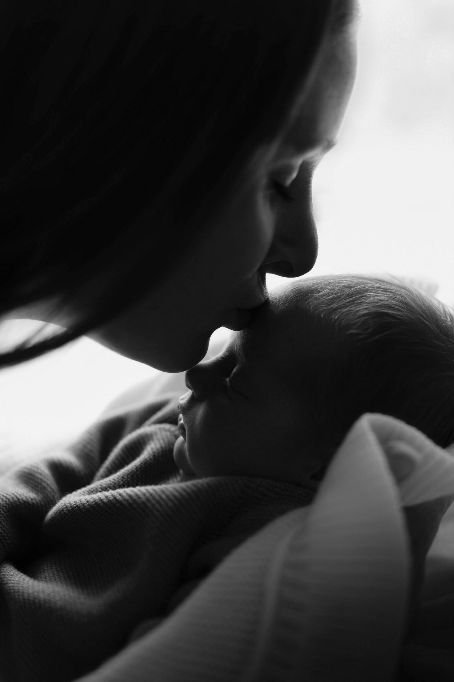 Black and white photo of a mother gently kissing her newborn baby, a tender and intimate moment of love.