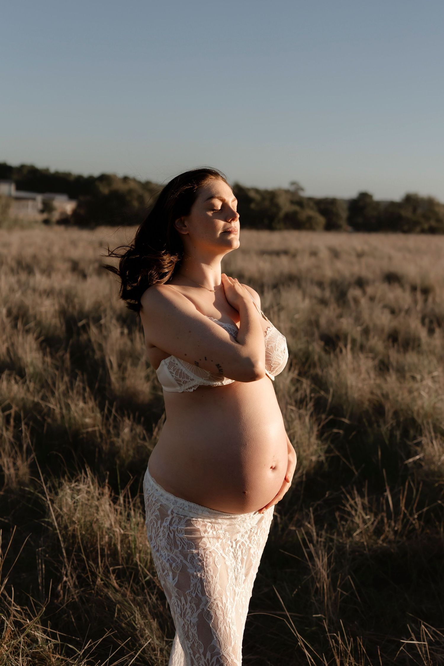 Pregnant woman in lace dress standing in golden field at sunset, cradling baby bump with eyes closed.