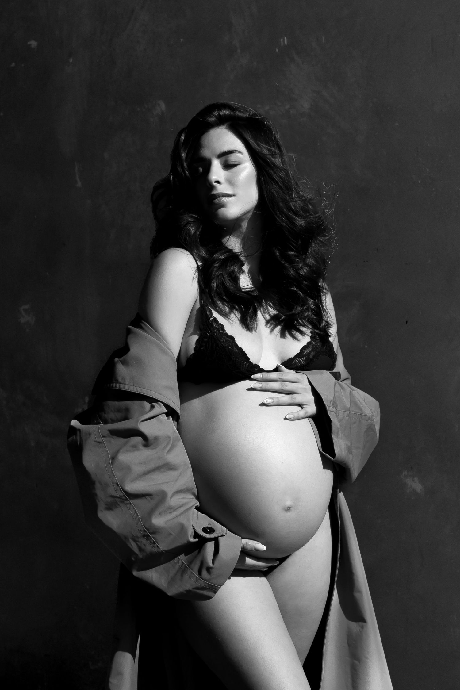 Black and white maternity photo of a pregnant woman in lingerie and open coat, cradling her baby bump.
