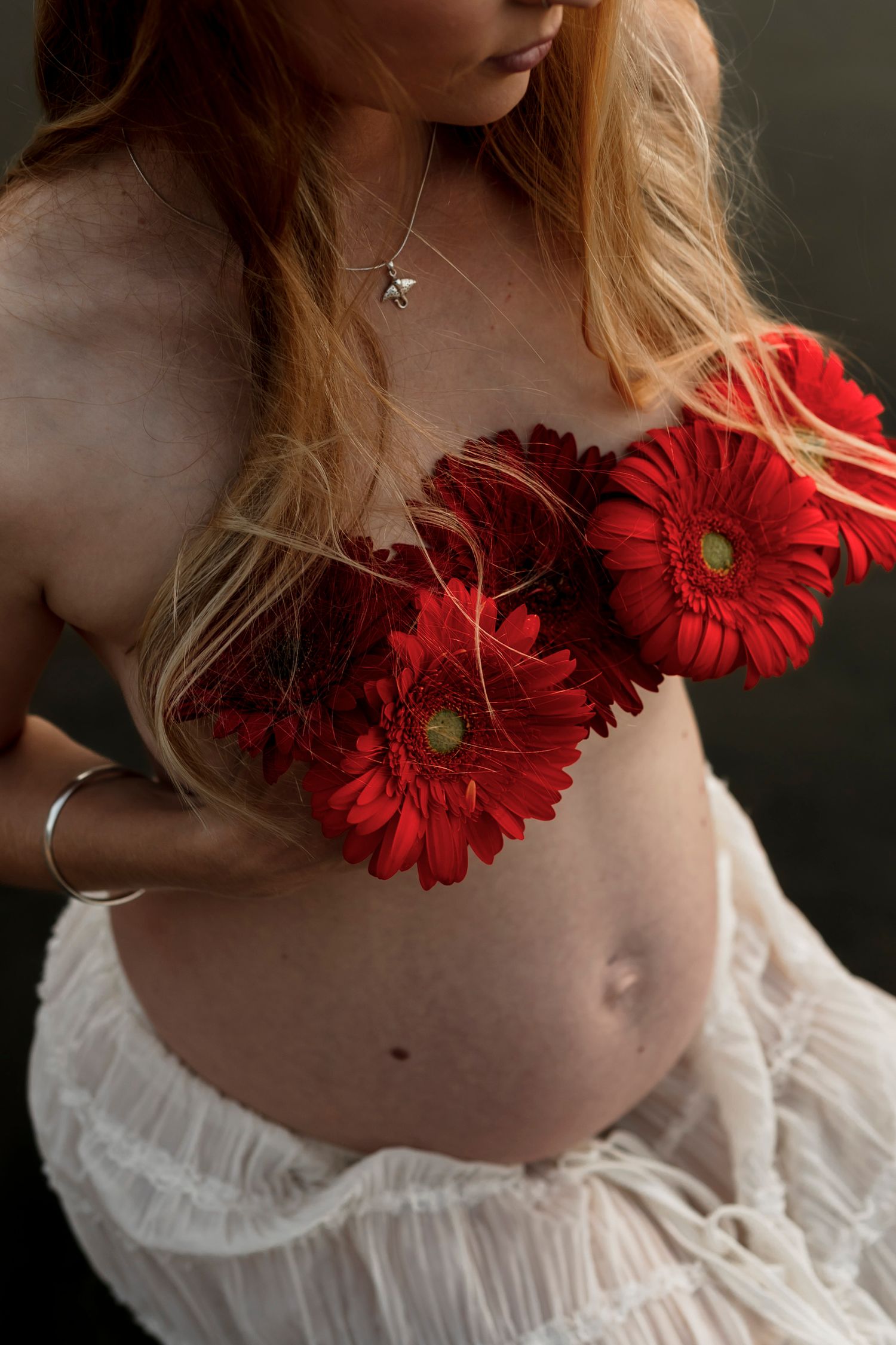 An artistic maternity photo showing a bare belly with vibrant red gerbera daisies and white flowing fabric.