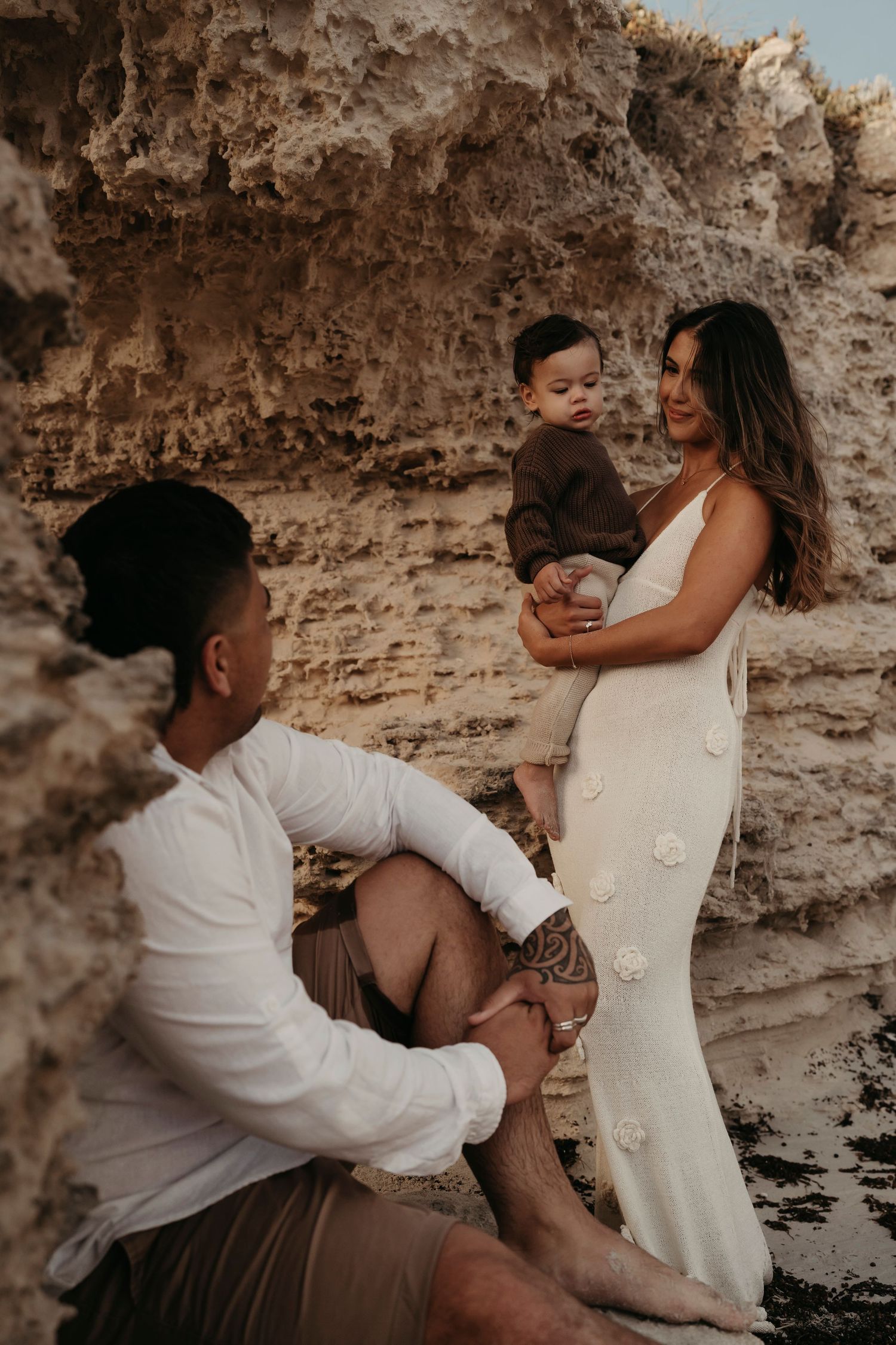 A family shares a tender moment together on a beach with rocky cliffs in the background during golden hour.