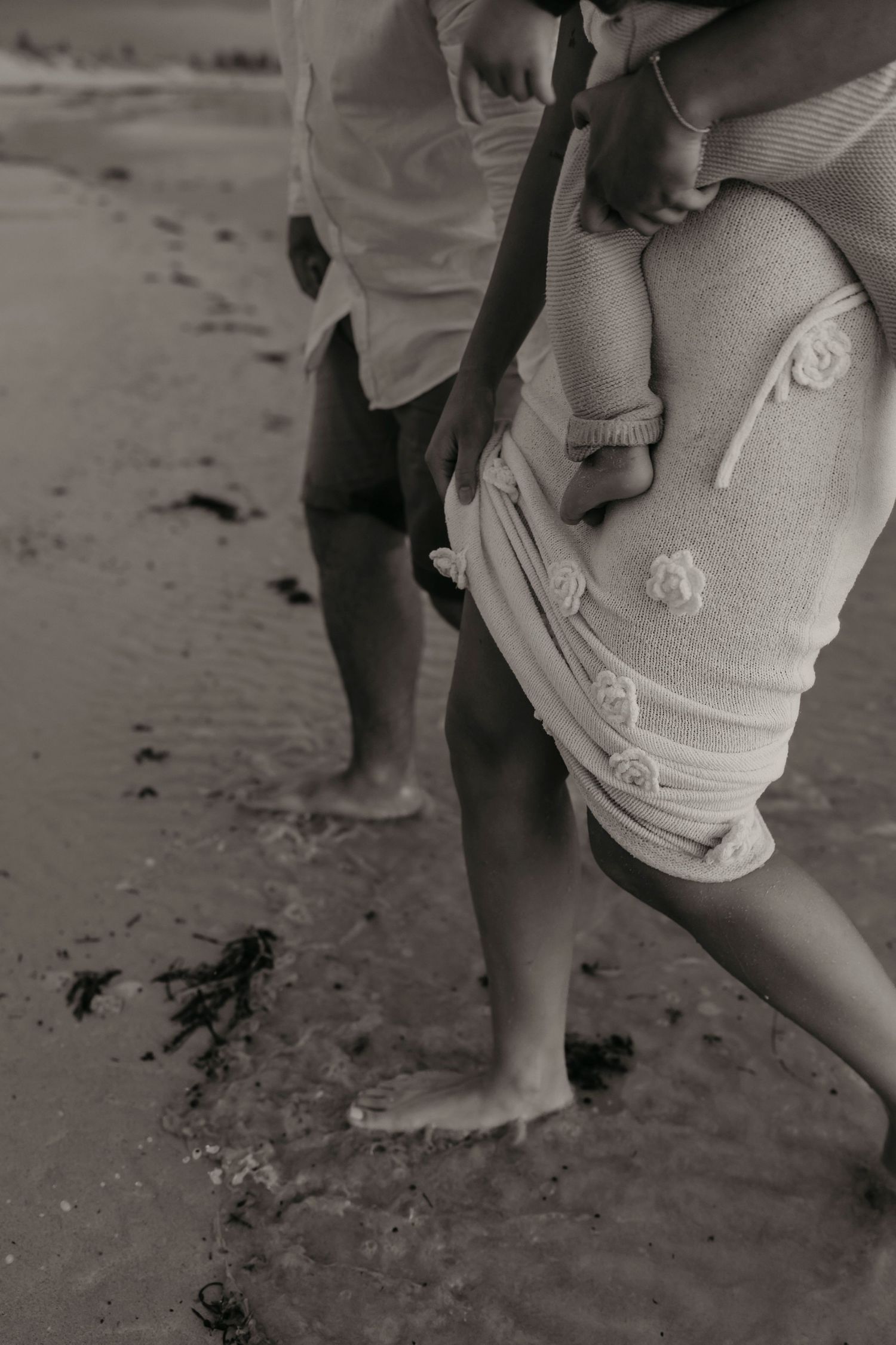 Black and white photo of bare feet walking on sandy beach with embroidered jeans visible.