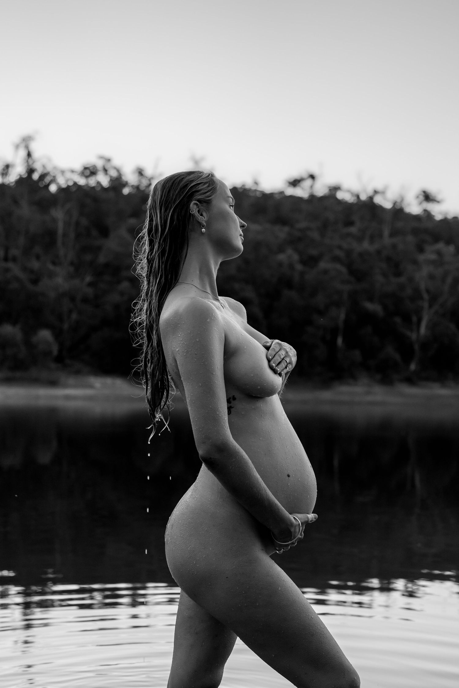 Monochrome maternity photo capturing a silhouette against a serene lakeside backdrop at dusk.