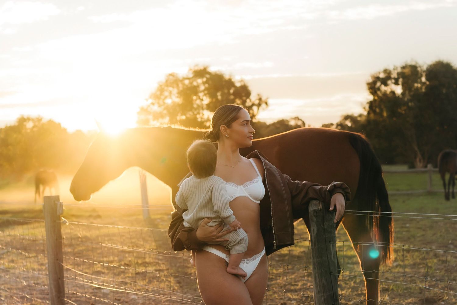 A couple embraces in a sunlit field with horses grazing in the background during golden hour.
