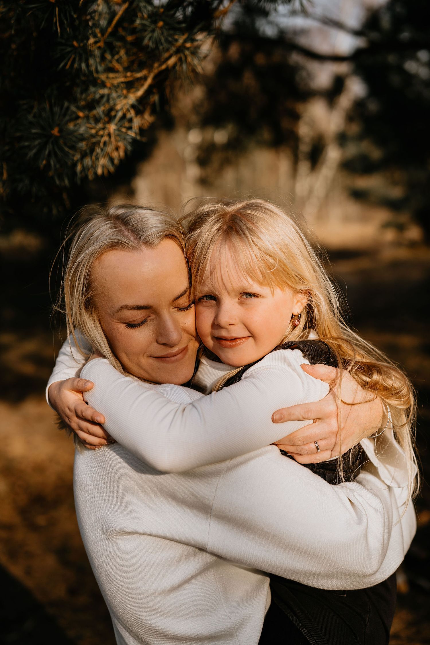 A tender embrace between parent and child outdoors during golden hour, both wearing white sweaters and smiling warmly.