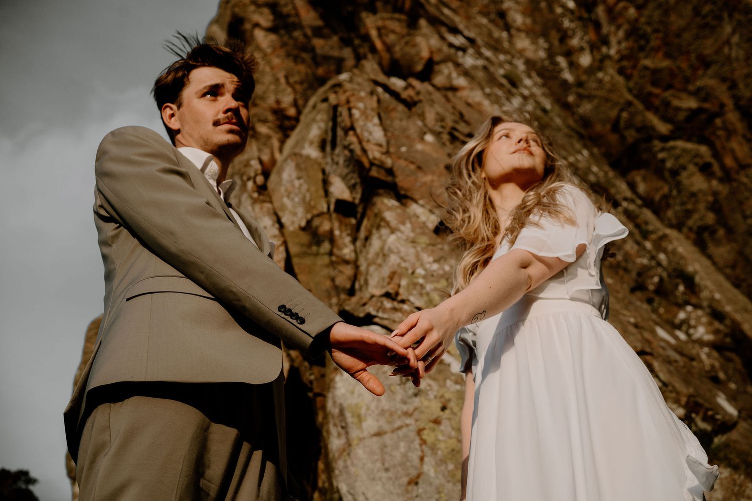 A couple in wedding attire holds hands in front of a dramatic rocky cliff during their outdoor ceremony.
